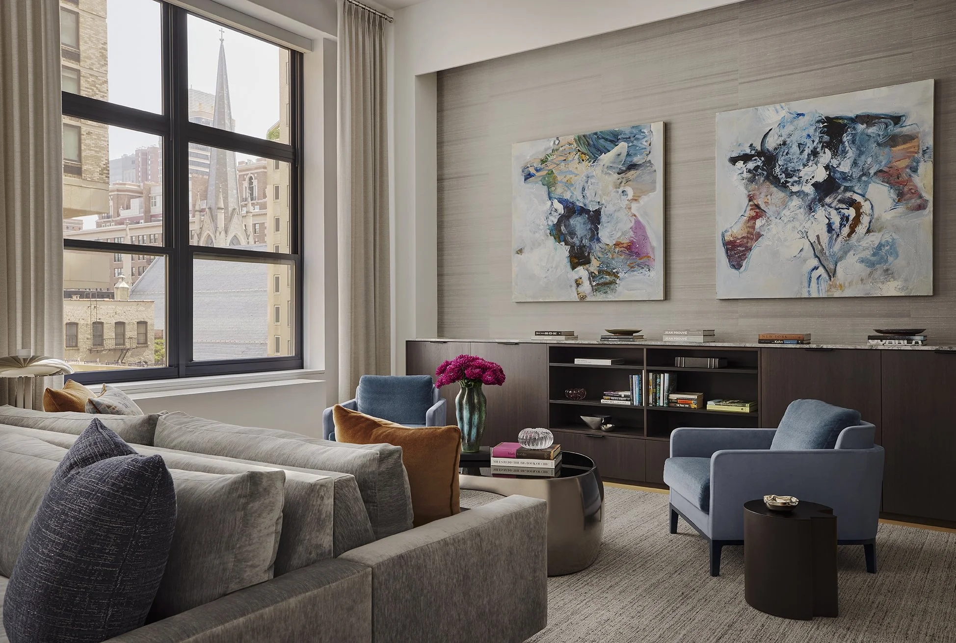 A contemporary living room with large windows showing city buildings and a church. The room has a beige sofa with dark cushions, a blue armchair, a coffee table with books and a flower vase, and abstract artwork on the wall.