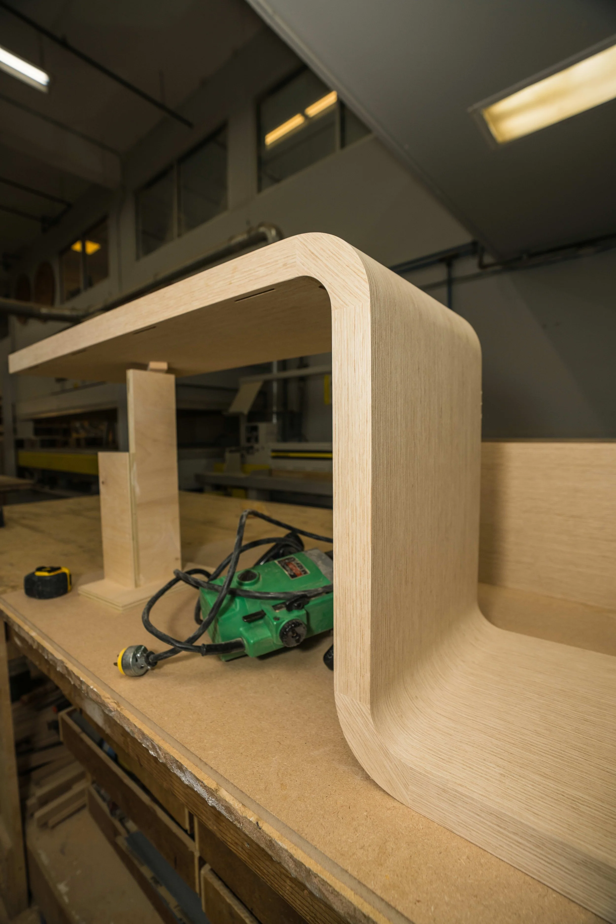 Wooden furniture piece in progress on a workbench, with tools including a green electric drill and a yellow measuring tape nearby, in a woodworking workshop.