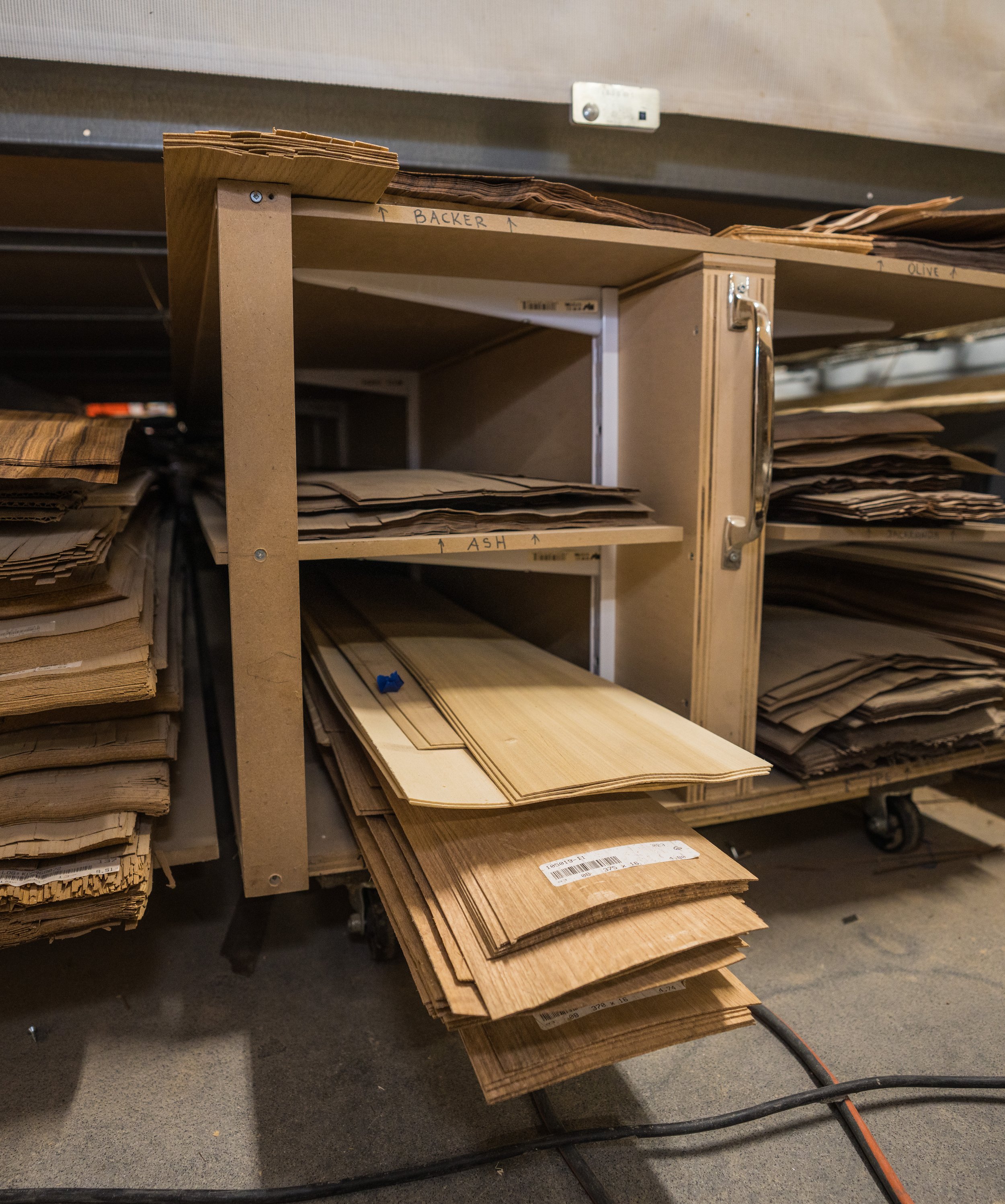 A wooden shelving unit with labeled shelves containing various wood and material samples in a workshop or storage area.
