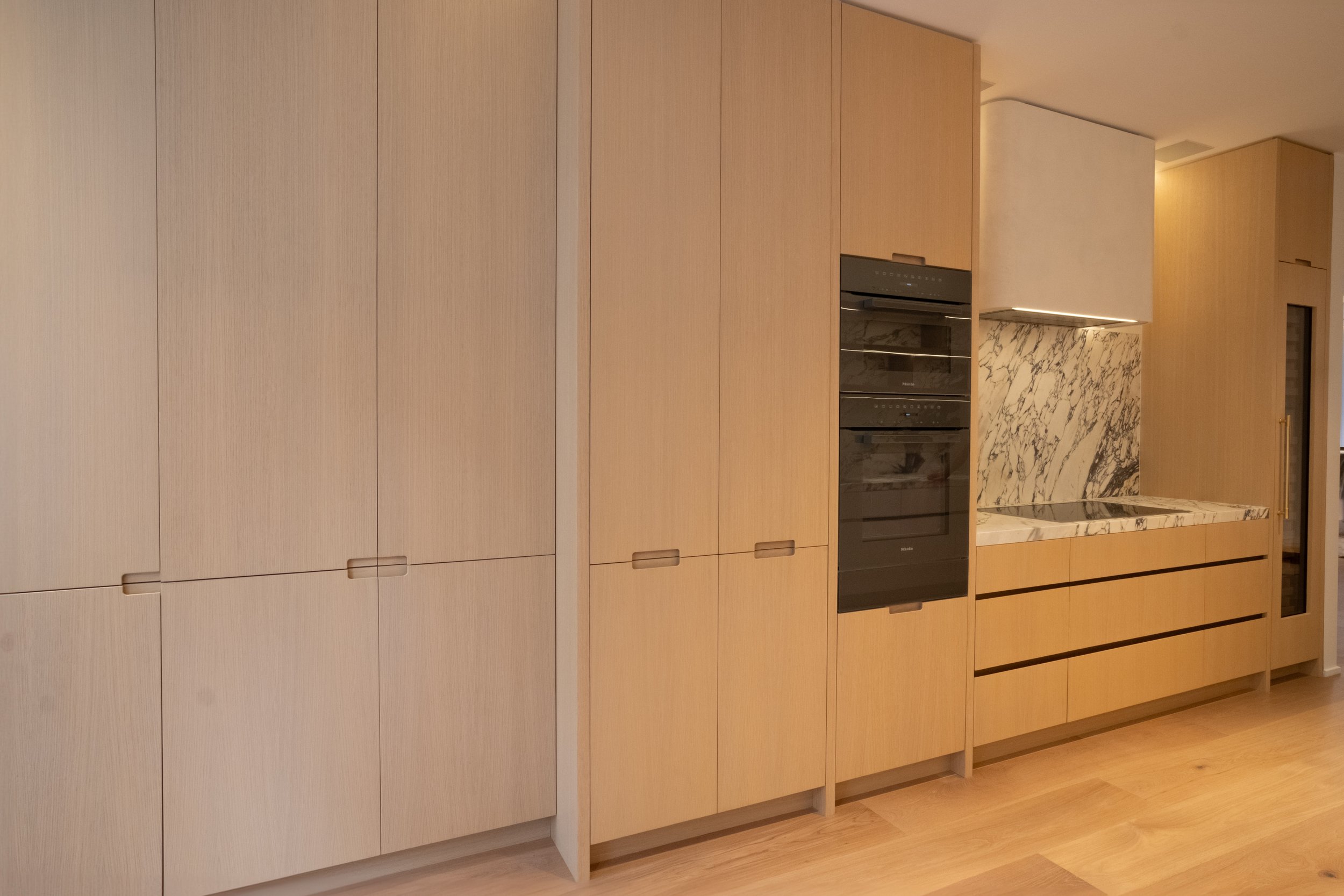 Modern kitchen with light wood cabinets, a marble backsplash, and built-in black oven appliances.