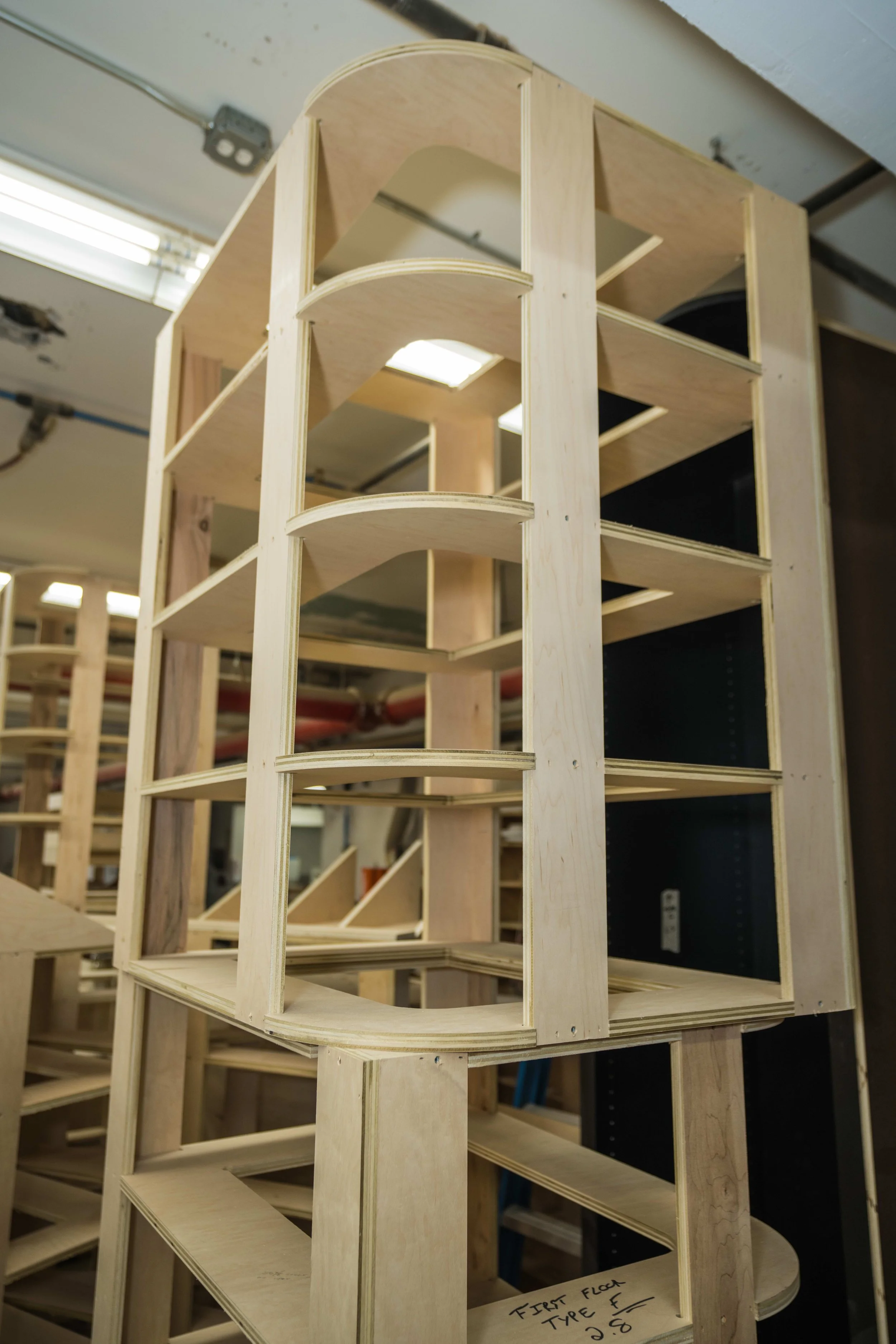 A wooden architectural model with multiple curved and straight shelves, labeled 'First Floor, Type F, 2.8', in a workshop setting.
