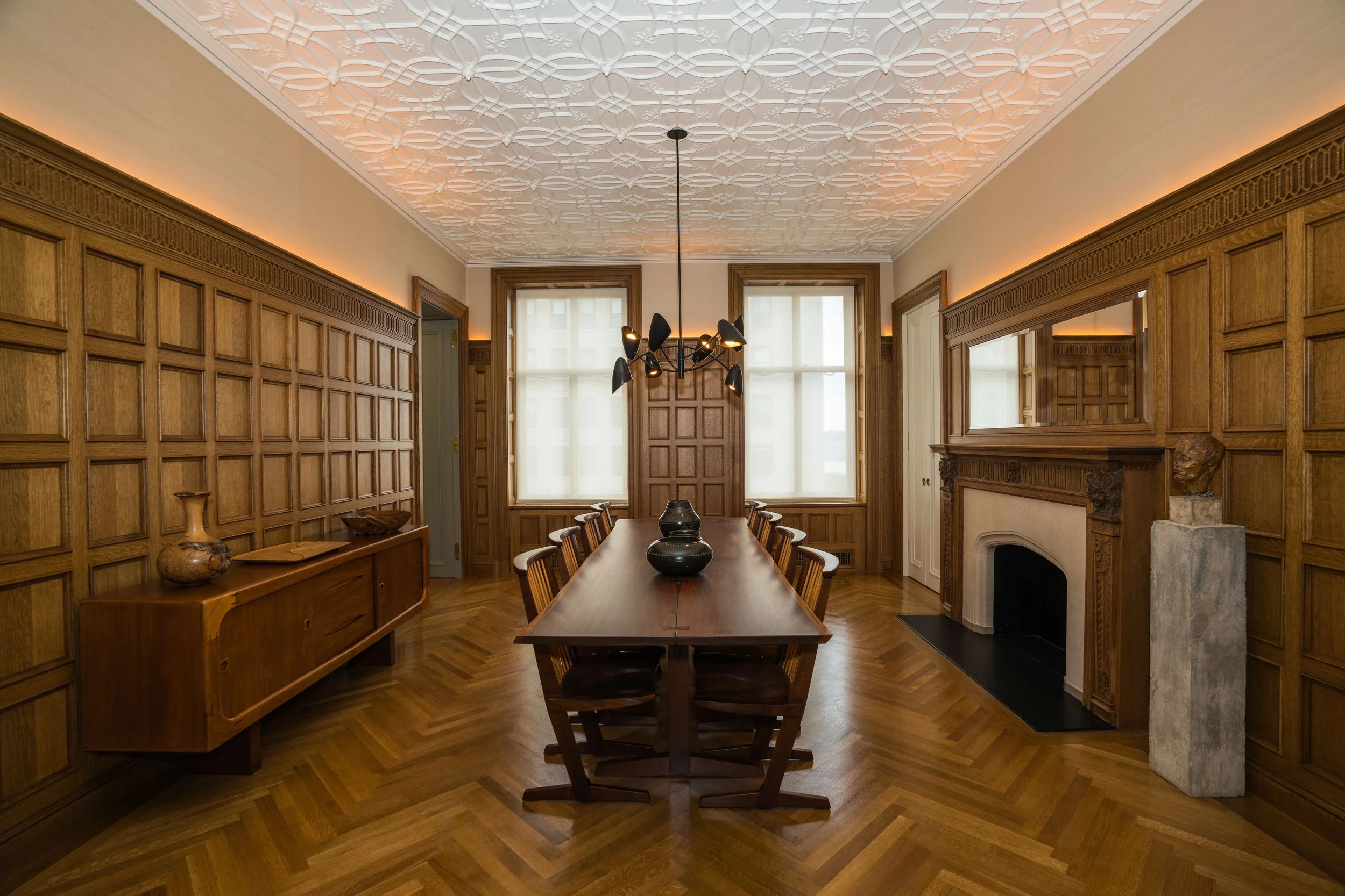 A room with wooden paneled walls, a wooden dining table with chairs, a black chandelier, large windows with blinds, and a fireplace with a detailed wooden mantel.
