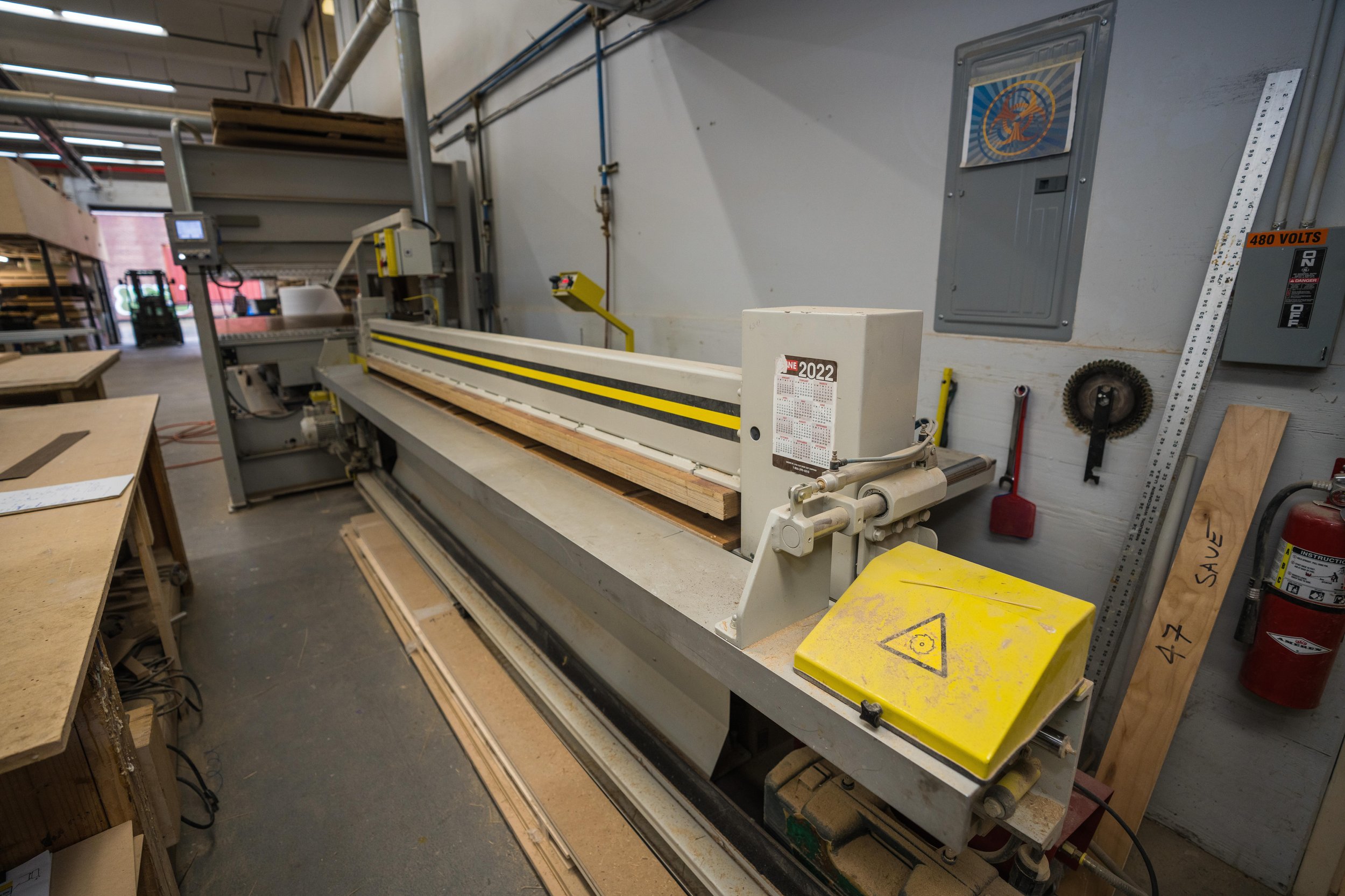 Large industrial saw machine in wood workshop, with wood pieces nearby and tools on the wall.