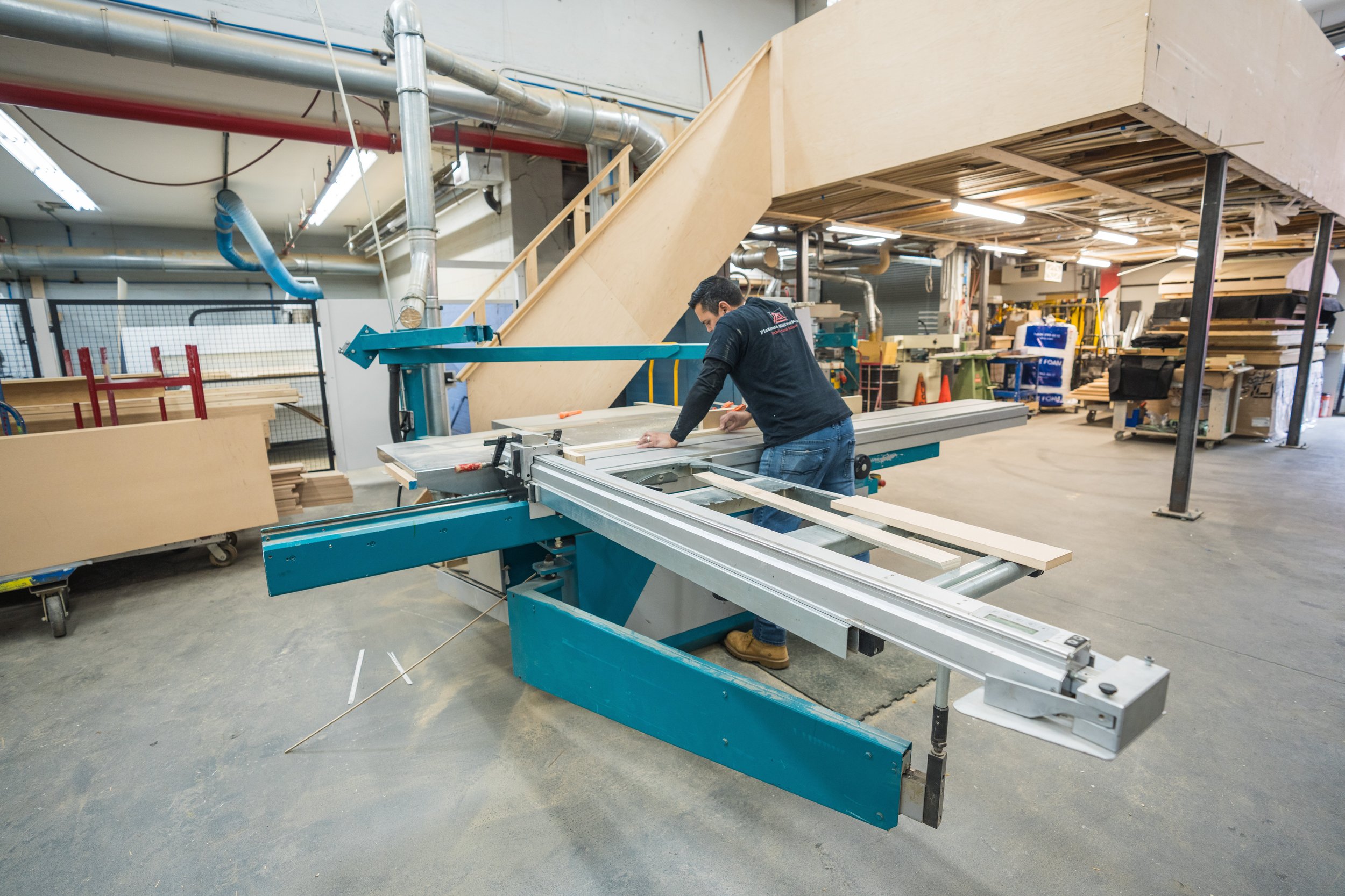 A man working in a woodworking shop, cutting or sanding a piece of wood on a large blue saw machine.