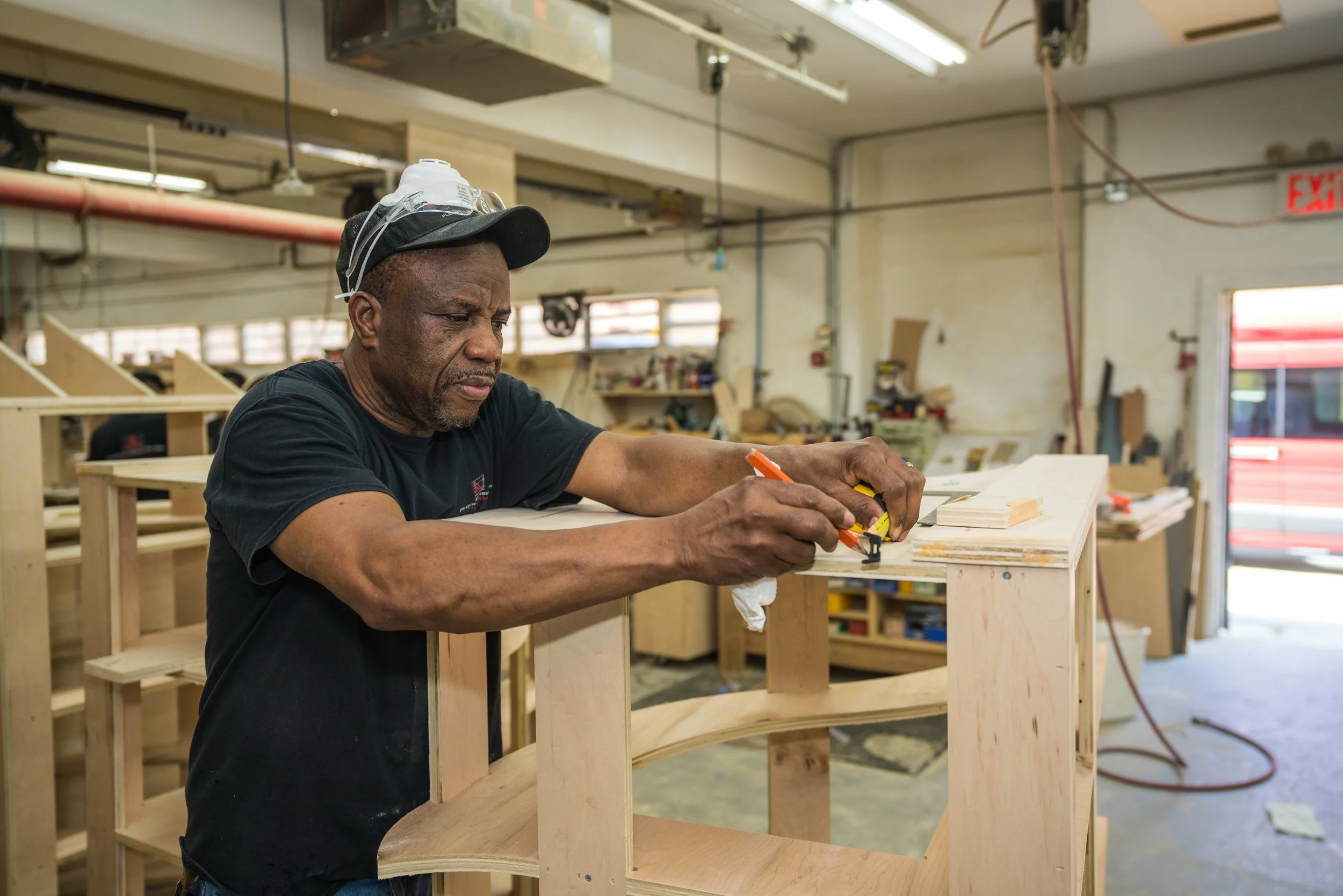 A man working on a woodworking project in a workshop, wearing a cap and safety goggles on his head, focused on measuring a piece of wood.