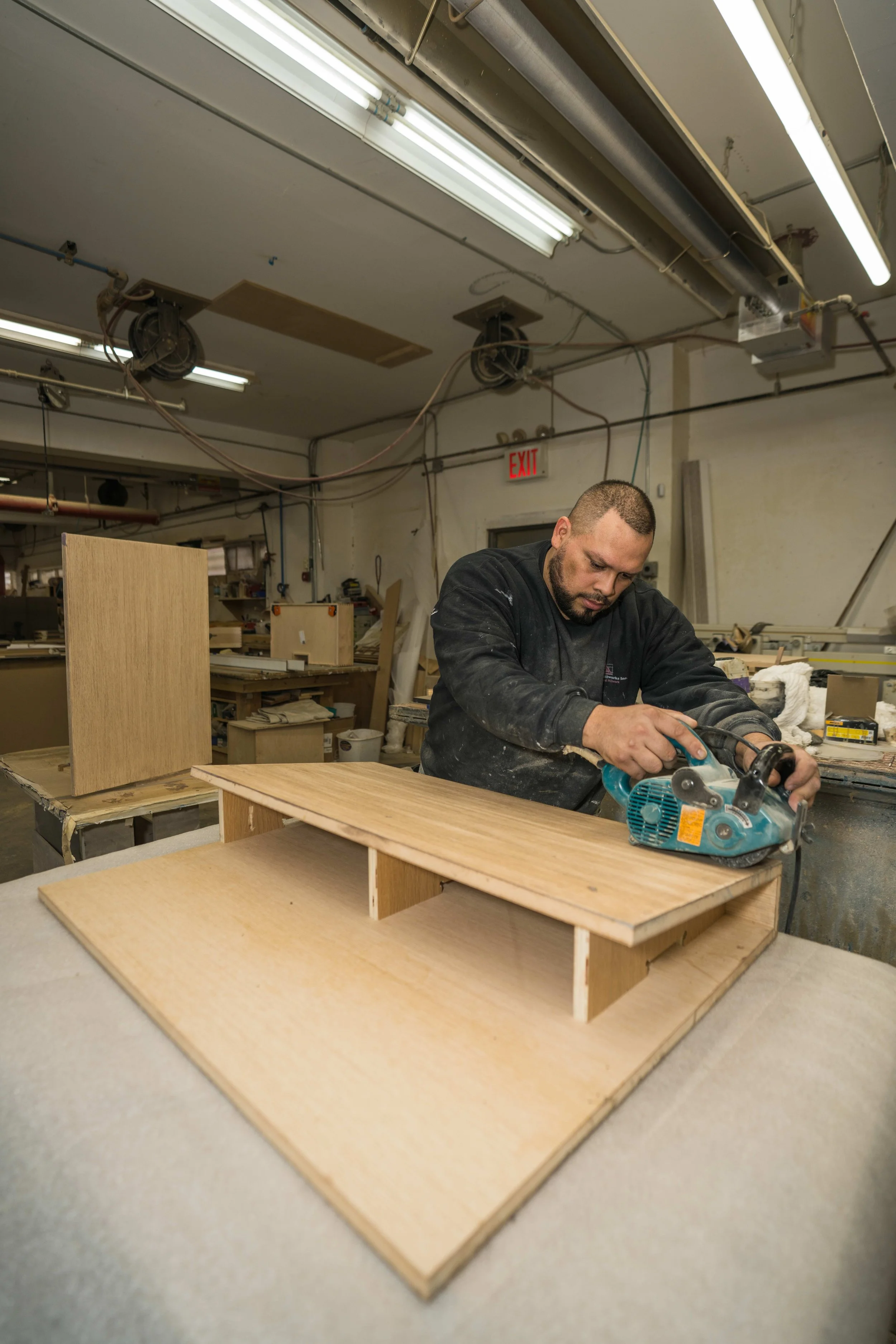 A man is working in a woodworking workshop, using a circular saw on a wooden project. Shelves with materials and tools are in the background, and an illuminated ceiling with fluorescent lights is overhead.