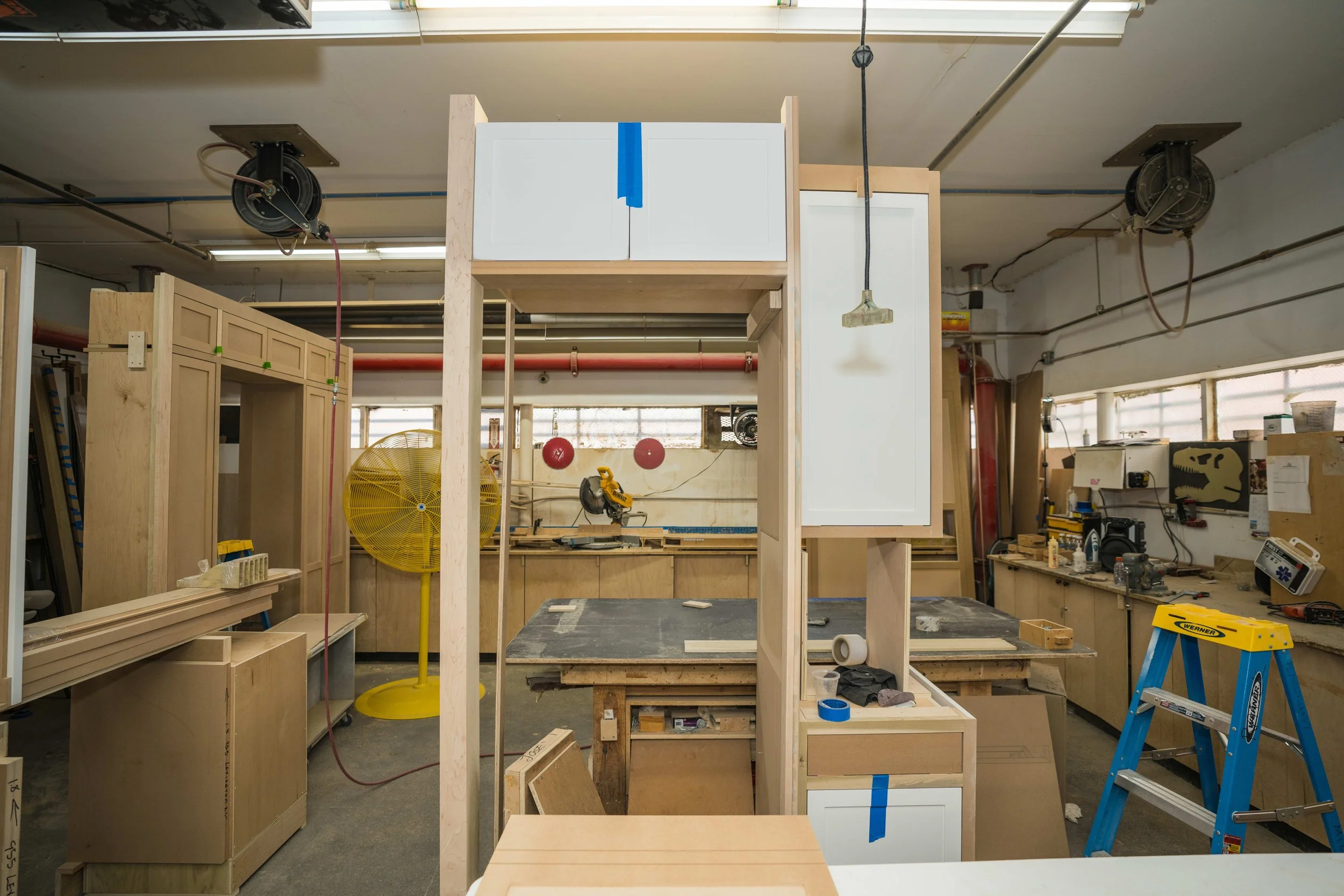 A woodworking workshop with unfinished wooden cabinets and furniture frames, tools, and equipment scattered around. Two large industrial rollers are mounted on the ceiling, a yellow fan, and various supplies on workbenches.
