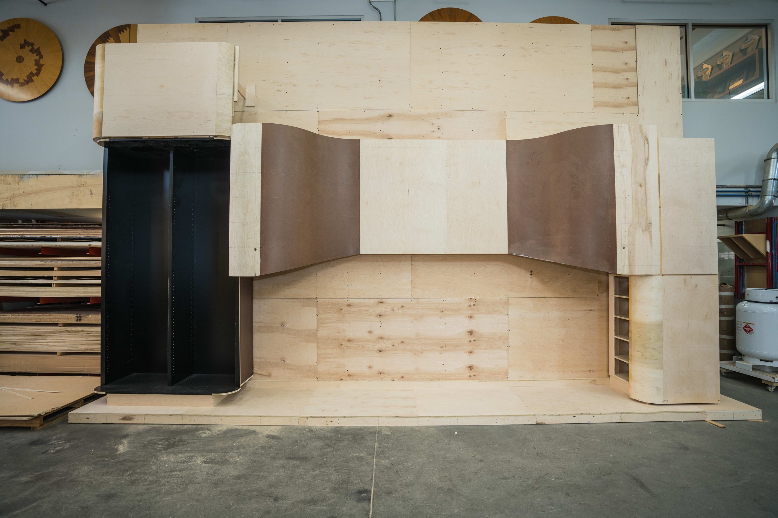 Wooden display structure in a workshop with circular wall decorations, storage shelves, and a propane tank in the background.