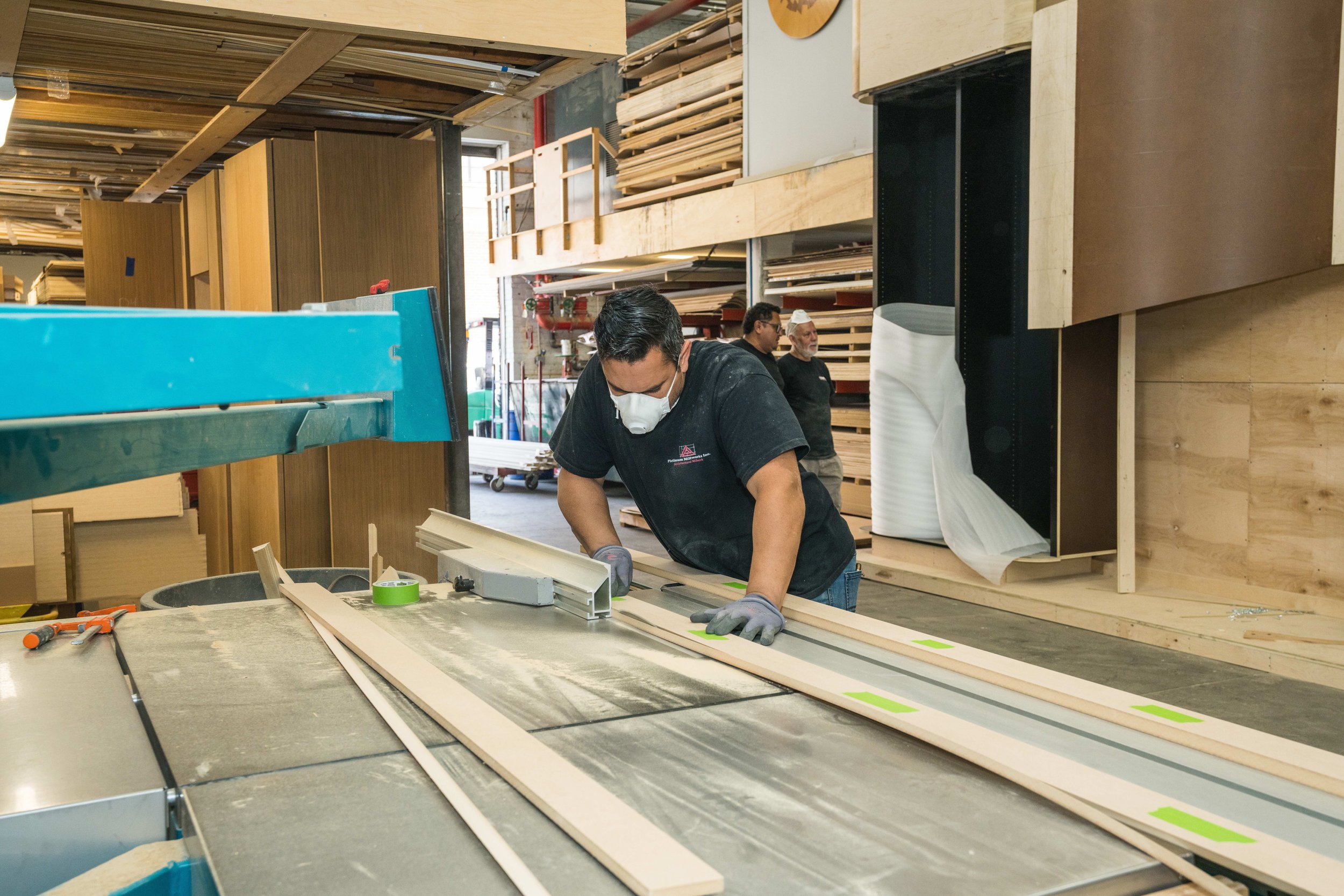People working in a woodworking shop with wooden planks and tools, one person wearing a mask and gloves, focused on a woodworking project.