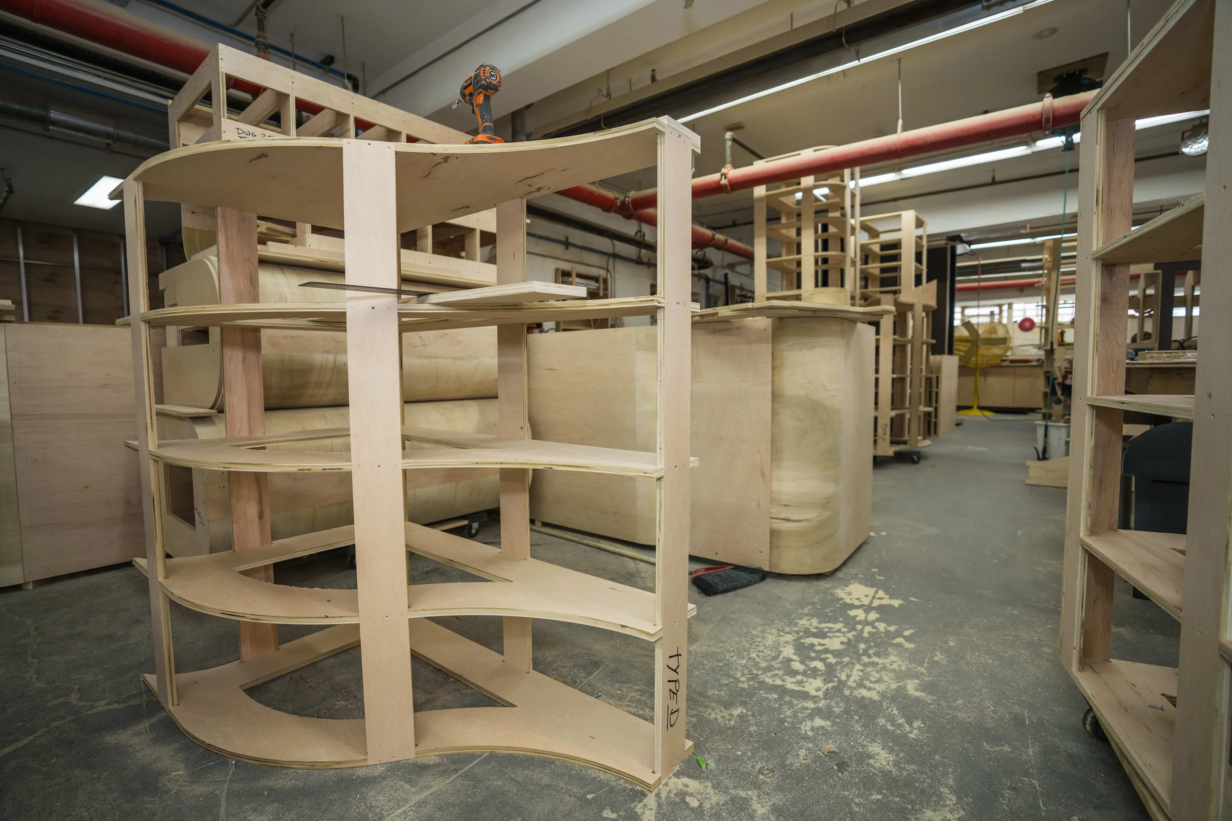 Wooden furniture pieces and shelves in a workshop, with tools and equipment, and unfinished wooden structures on the floor.