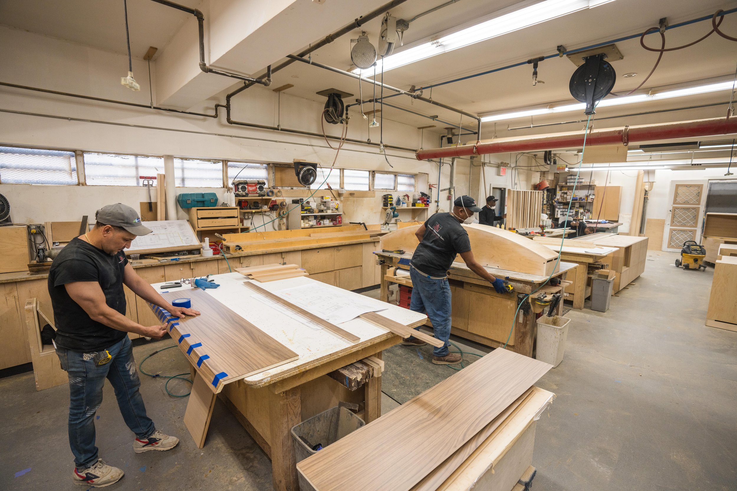Three workers in a woodworking shop working on large wooden furniture pieces surrounded by various tools and materials.