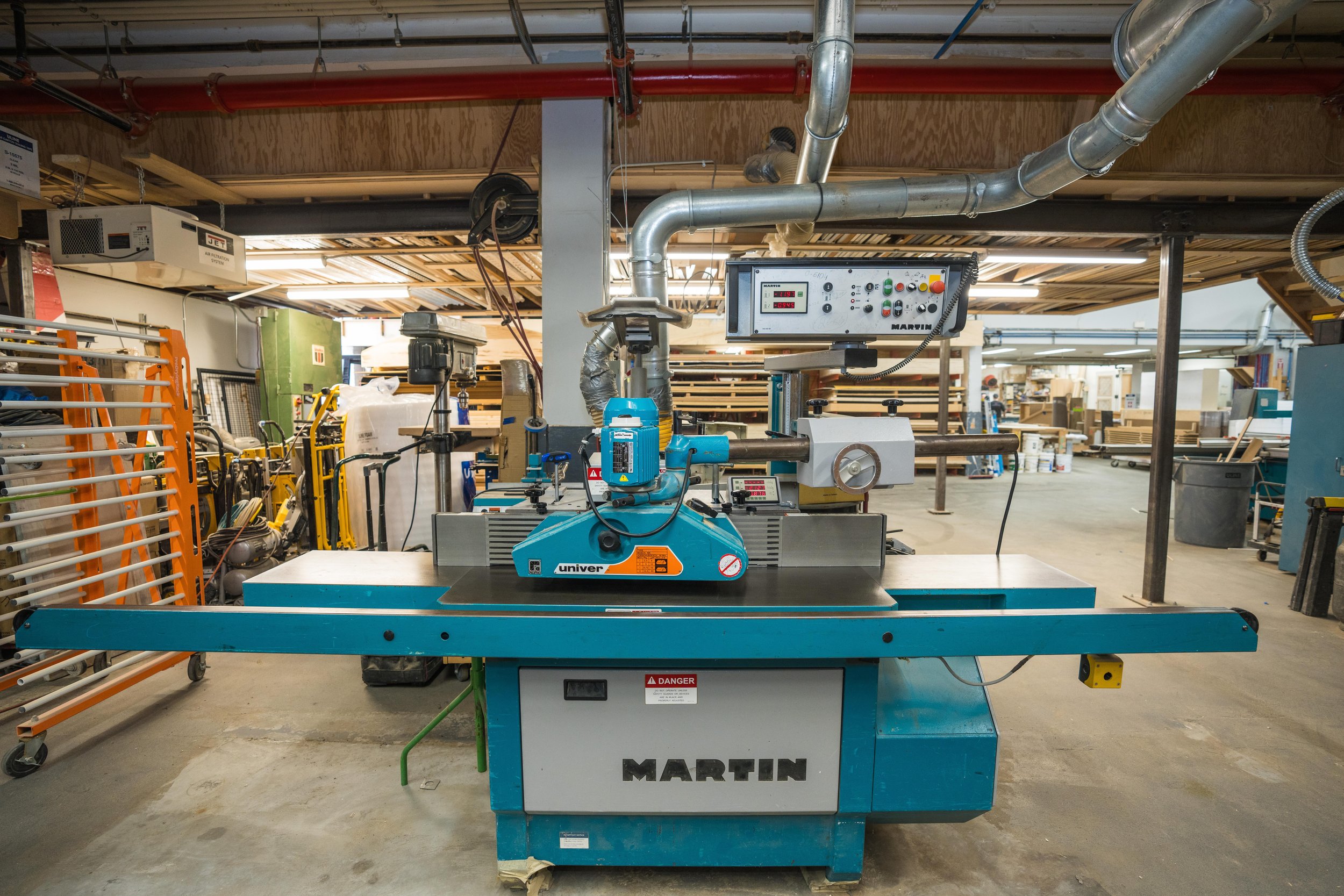 Industrial woodworking machine in a workshop, with a blue base labeled 'Martin' and various control panels, pipes, and other equipment around it.