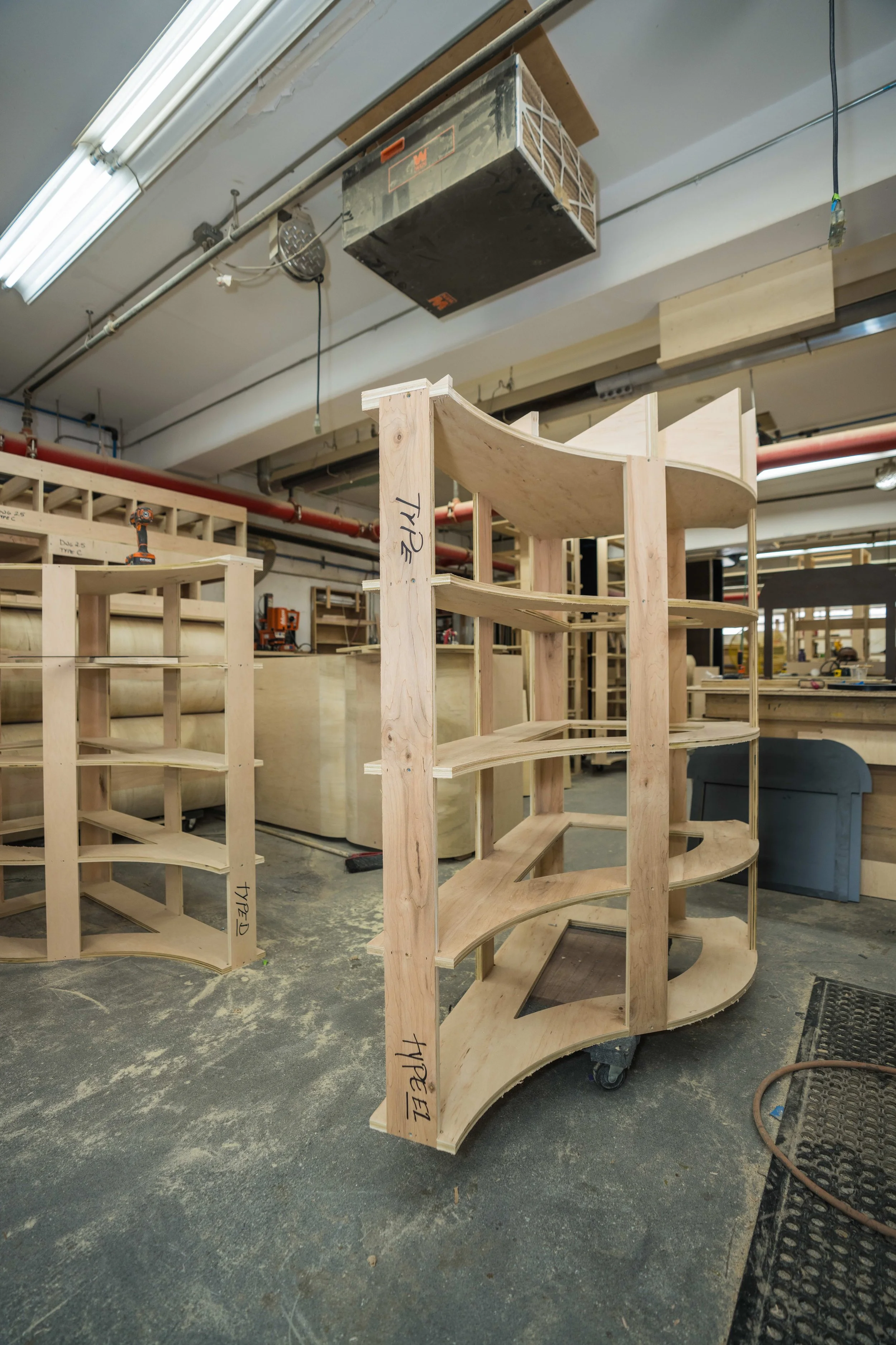 Wooden curved shelves or display units in a woodworking workshop, with some parts labeled with handwriting.