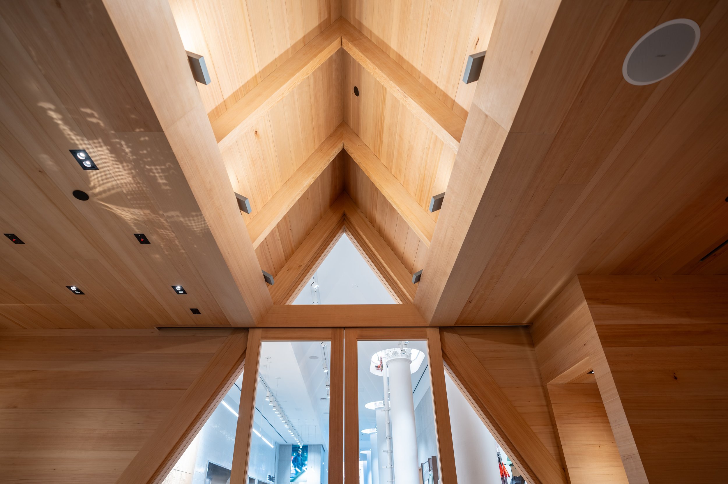 View looking up at a wooden ceiling with triangular cutouts and glass windows, with a white column and parts of a modern interior space visible through the openings.