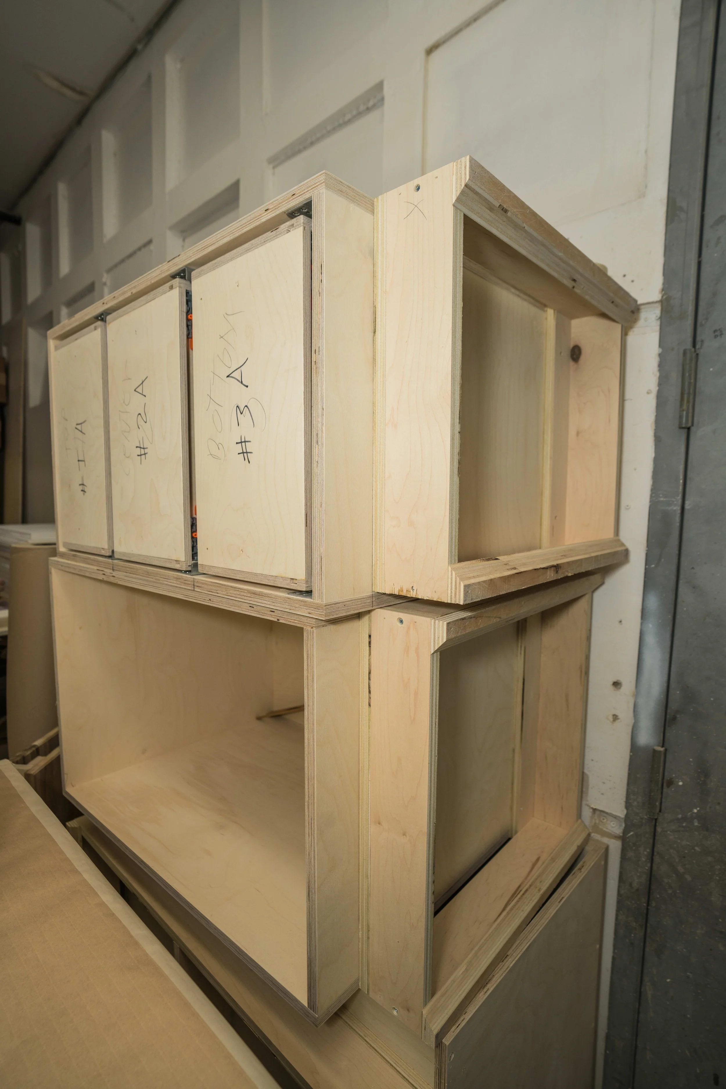 Wooden shelf units with multiple open compartments, some marked with handwritten notes, assembled in a workshop or storage area.