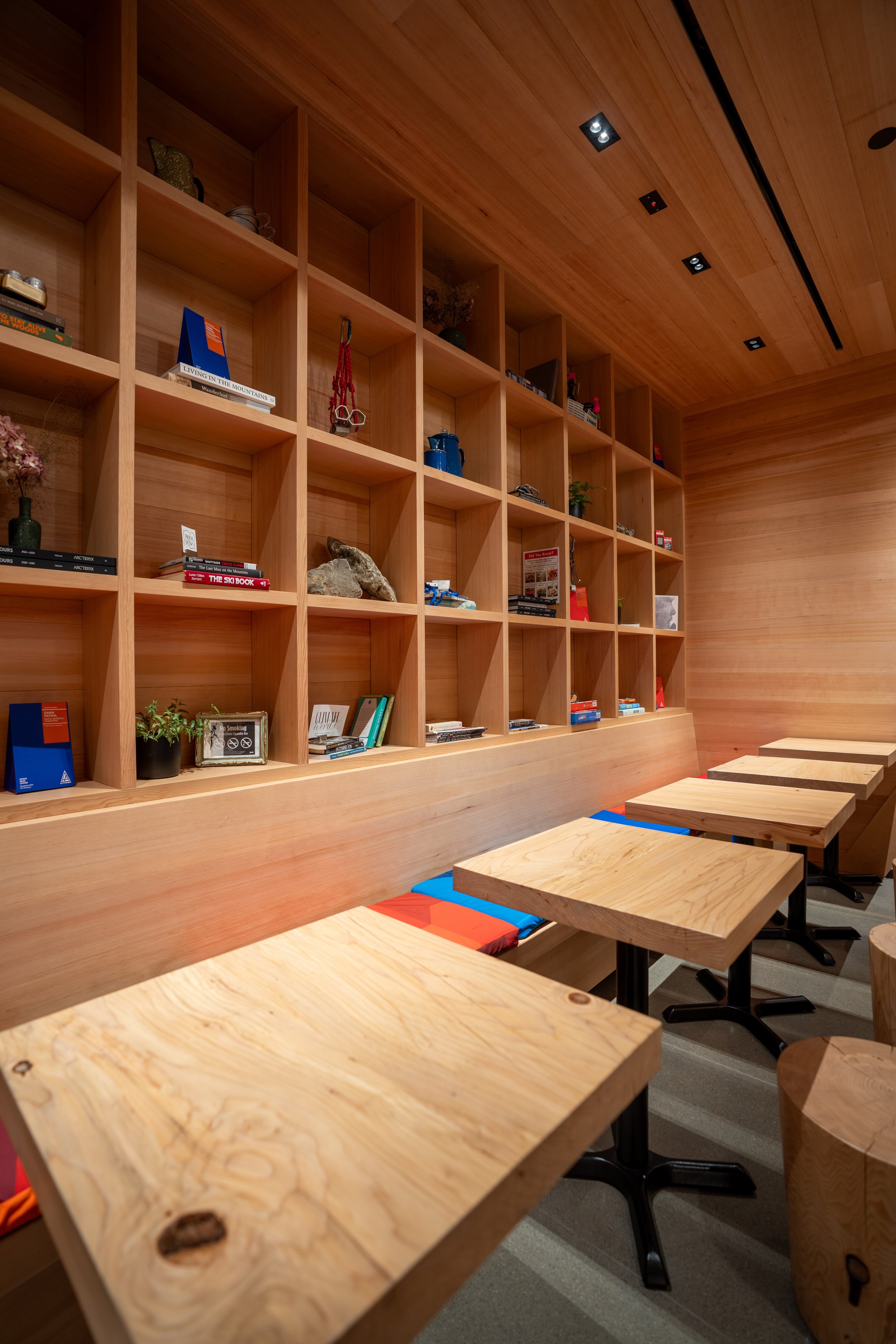 Wooden shelf wall with books and decorative items, five wooden tables with black bases and seat cushions in blue and orange, in a modern interior space.