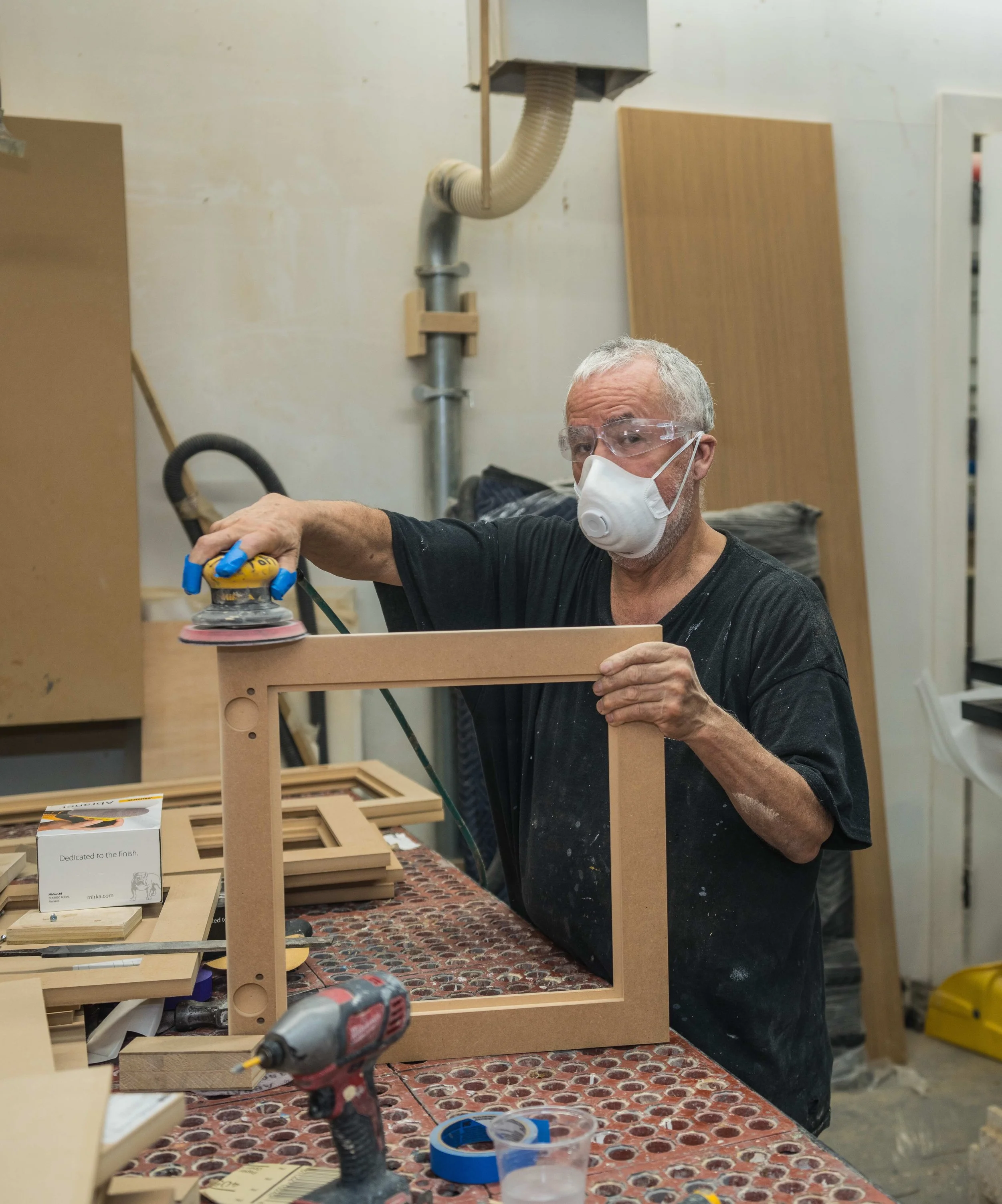 A man with gray hair, glasses, and a face mask is working on a woodworking project in a workshop. He is using a power sander on a wooden frame or piece, with various tools and materials on the workbench in front of him.