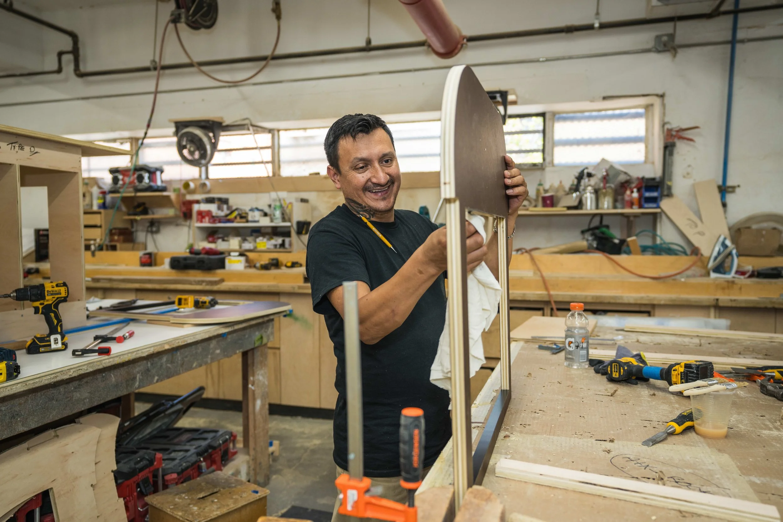 A man smiling while working on a woodworking project in a workshop, holding a piece of wood and surrounded by tools and workshop supplies.