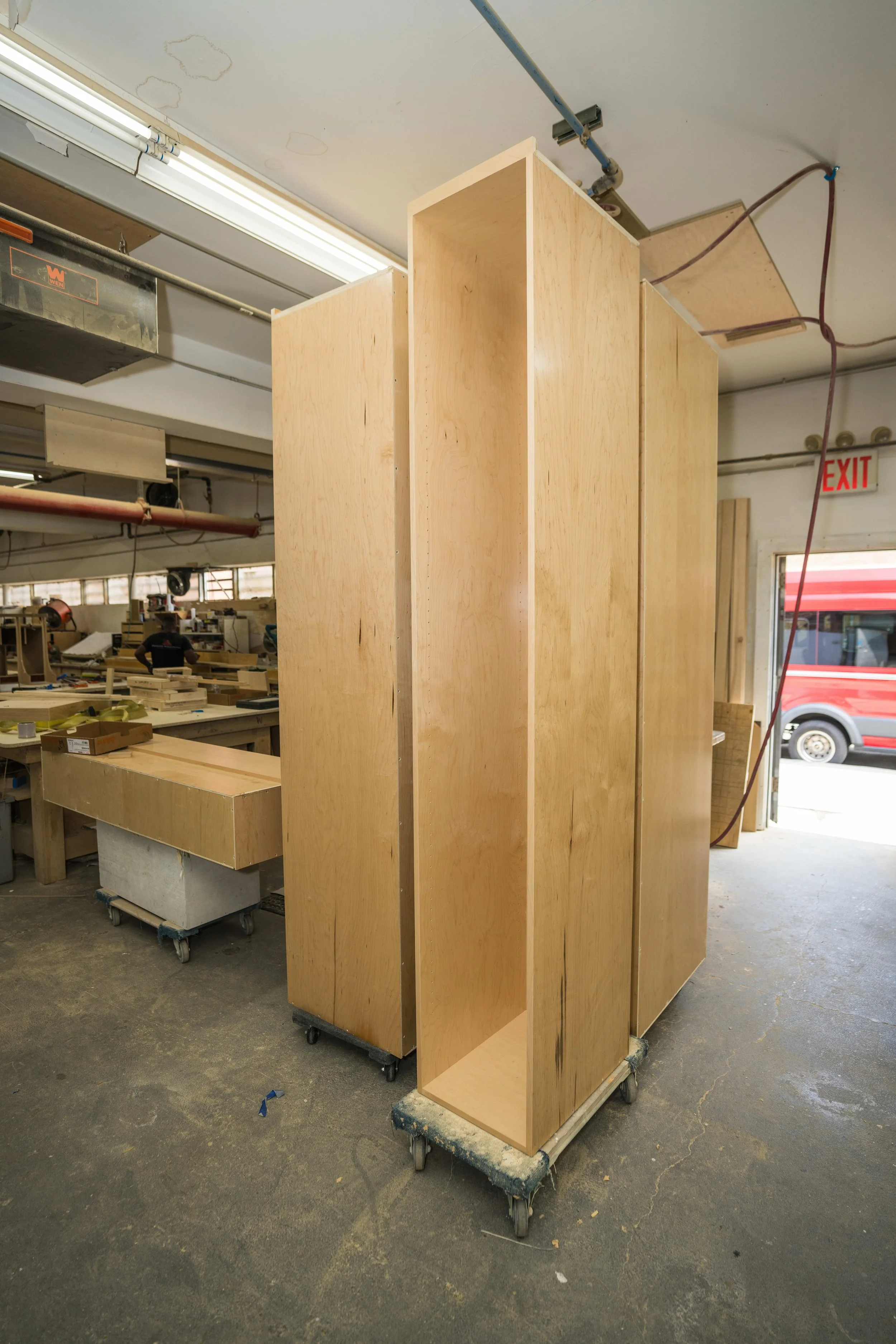 Workshop scene with unfinished wooden cabinets on casters, workbenches, tools, and a person working in the background. Fluorescent lights and plumbing pipes are visible on the ceiling, with an exit sign near a door leading outside.