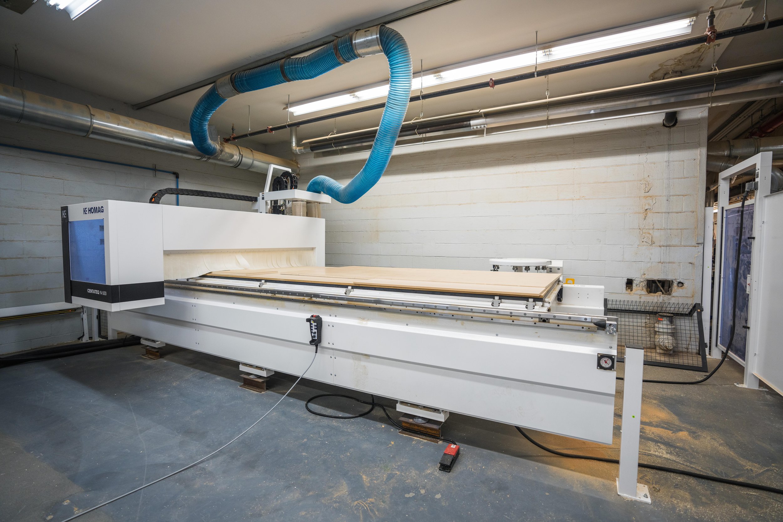 Large CNC machine manufacturing wood panels in a workshop with blue ducting and cinder block walls.