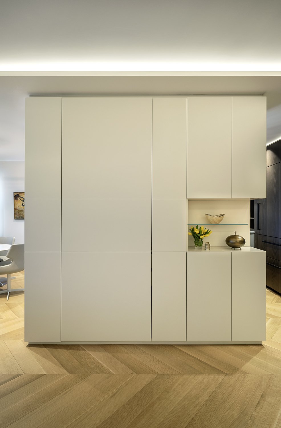 A modern white cabinet with decorative items and a glass shelf inside an open compartment, positioned in a contemporary home with light wood flooring.