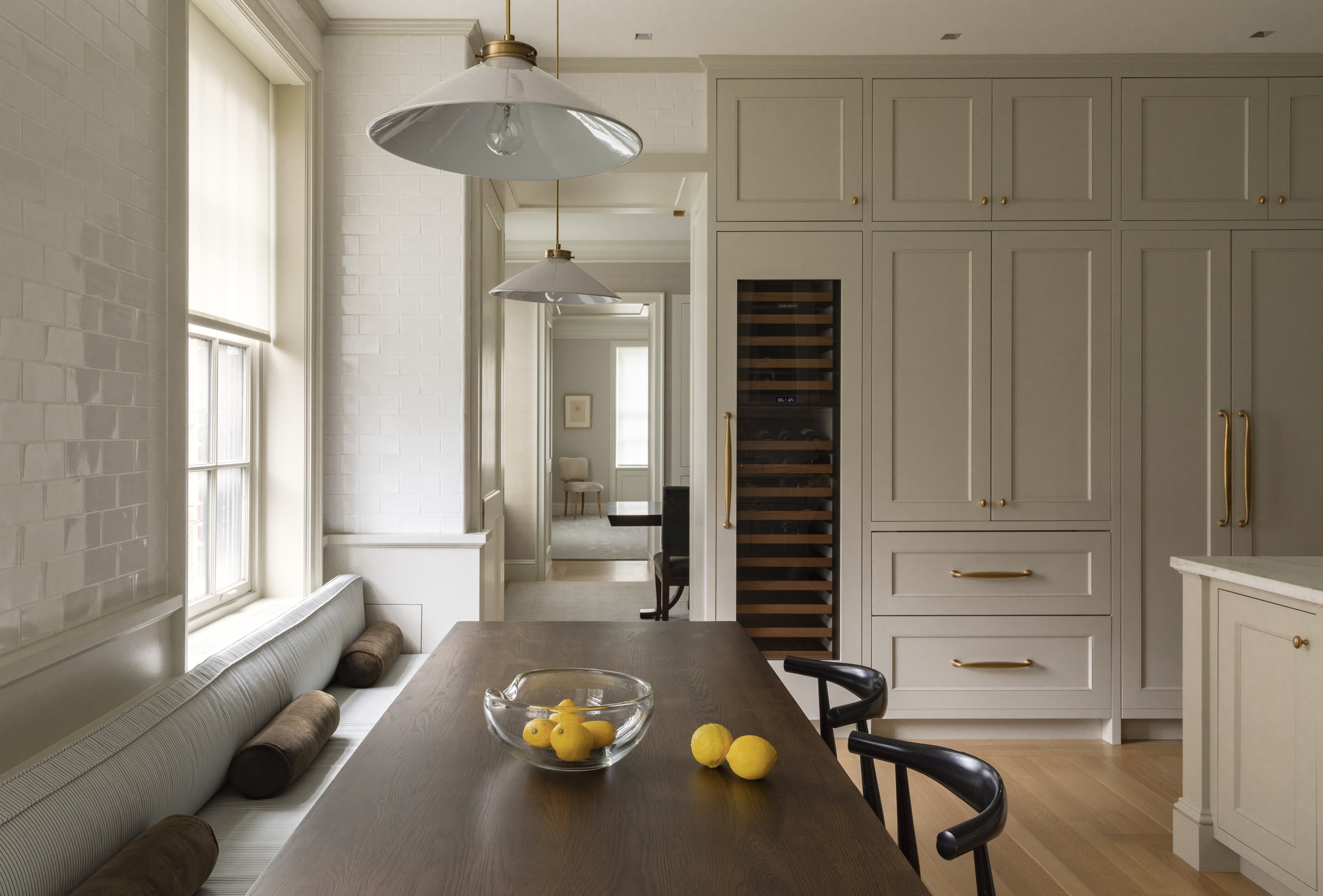 The image shows a dining area with a wooden table, black chairs, a built-in bench with brown cushions, and a grey kitchen cabinet with gold handles. There is a bowl of lemons on the table and two hanging white pendant lights. Natural light streams in