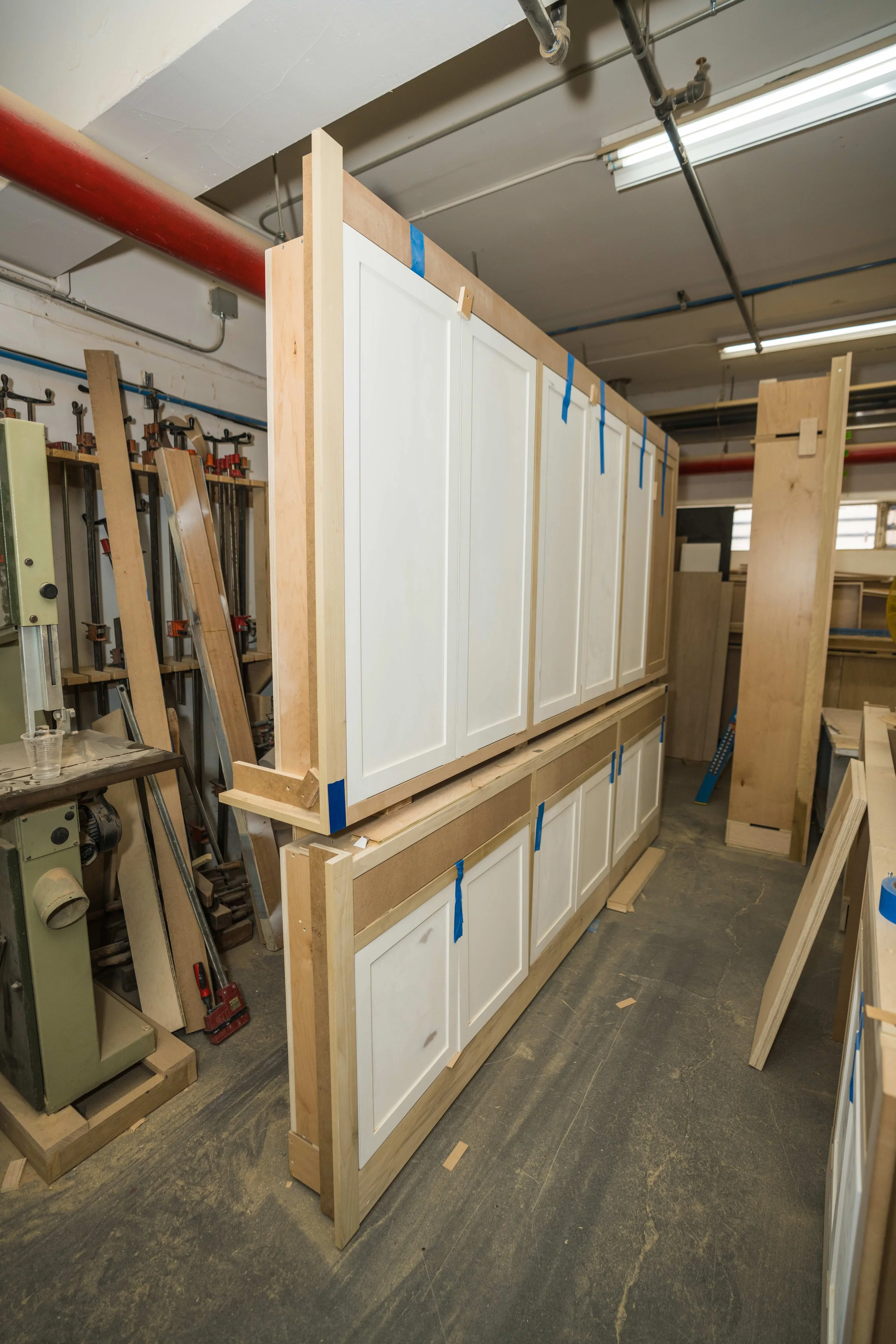 Wooden kitchen cabinets with white doors being assembled in a workshop, with clamps and woodworking tools visible around.