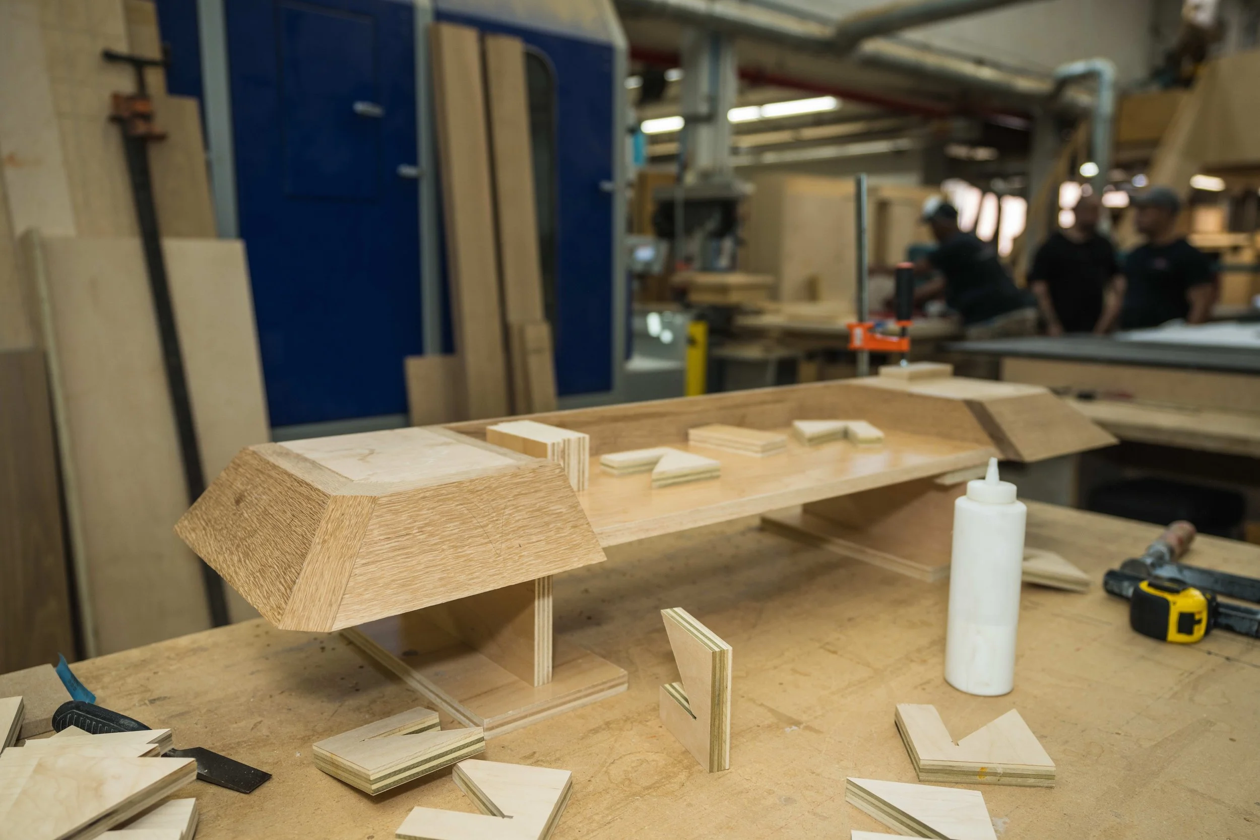 Woodworking workshop with a partially assembled wooden piece, clamps, glue, and scattered wood pieces on a workbench.