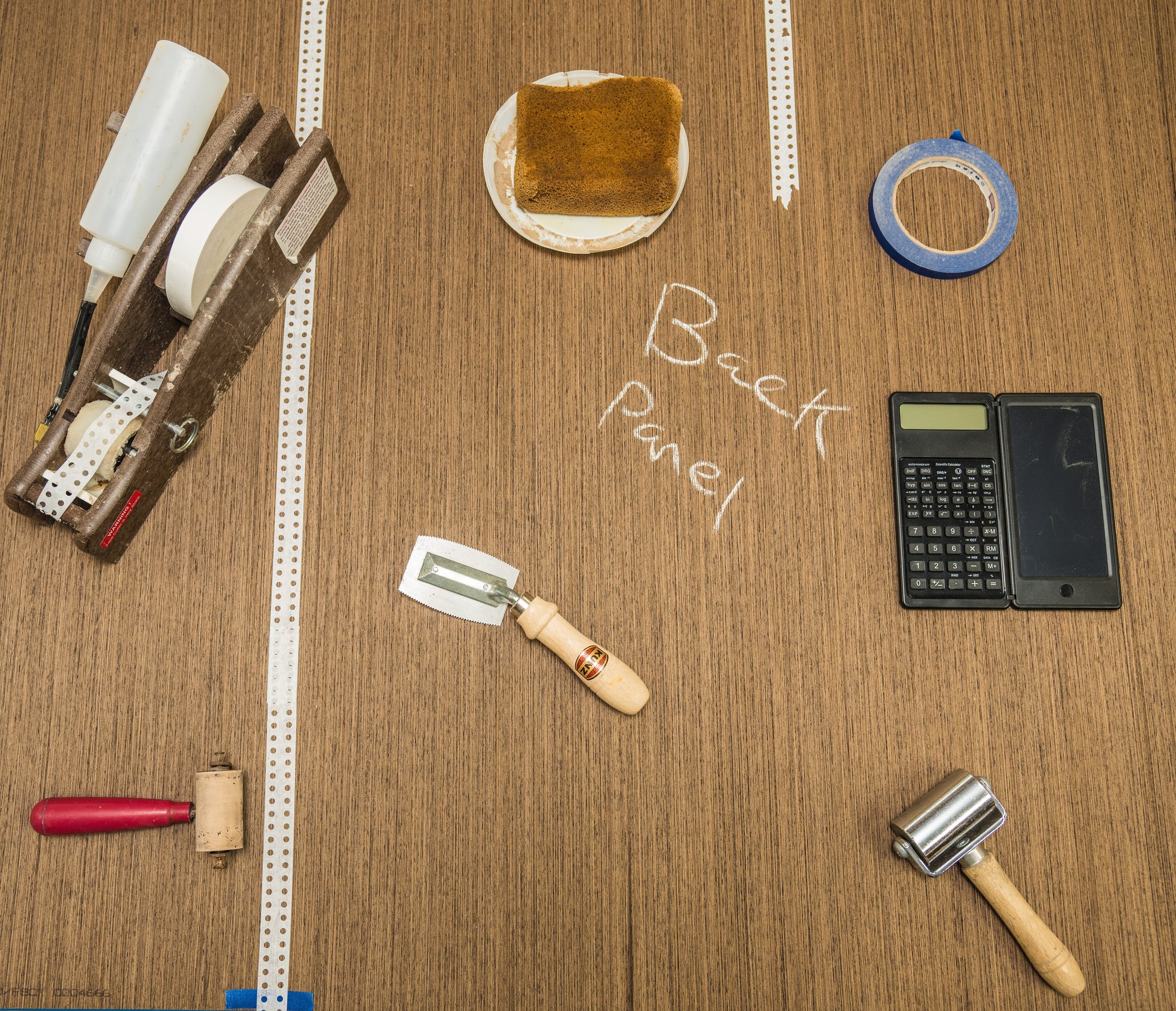 Craftsman workspace with tools and materials, including a woodworking glue, sanding sponge, measuring tape, small saw, calculator, and a hammer. 'Beck Panel' is written in chalk on the wooden surface.