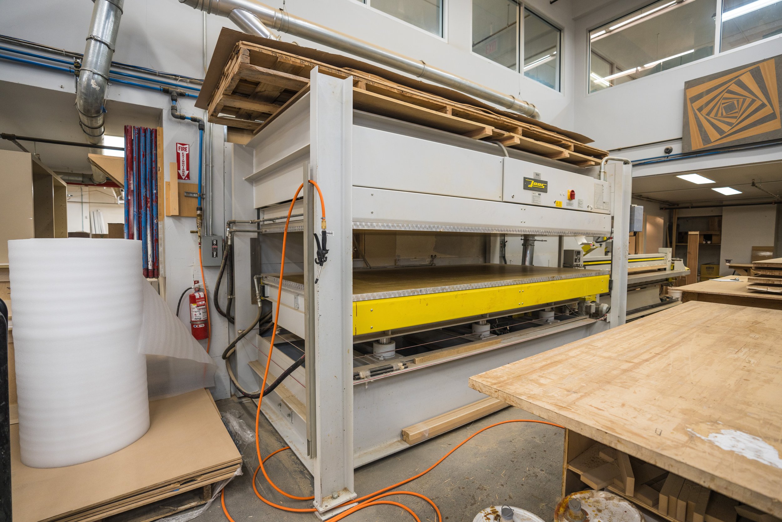 Industrial woodworking machine in a workshop surrounded by wooden sheets and tools.