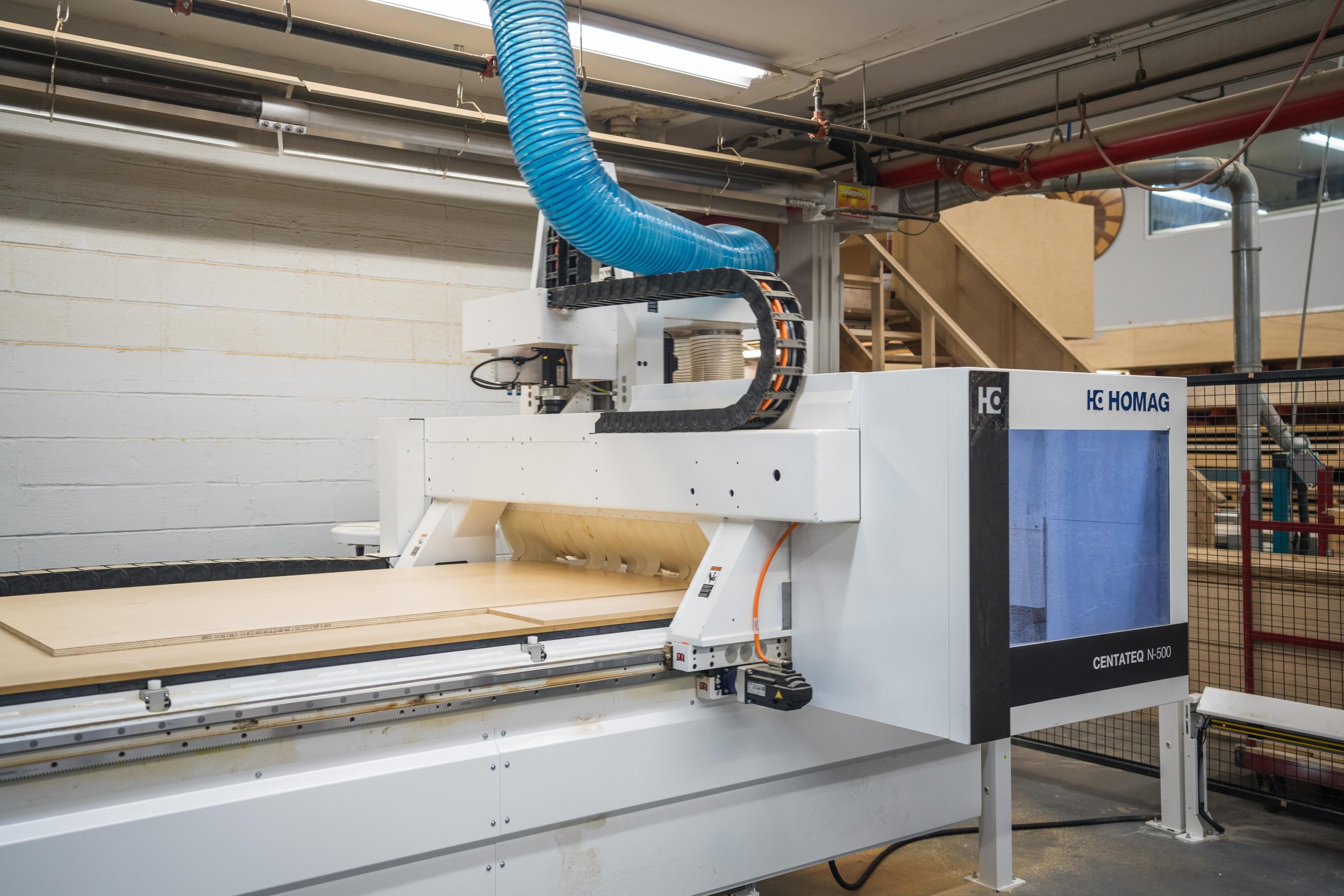 A CNC woodworking machine in a workshop, with a large blue exhaust hose above it, working on a wooden panel.