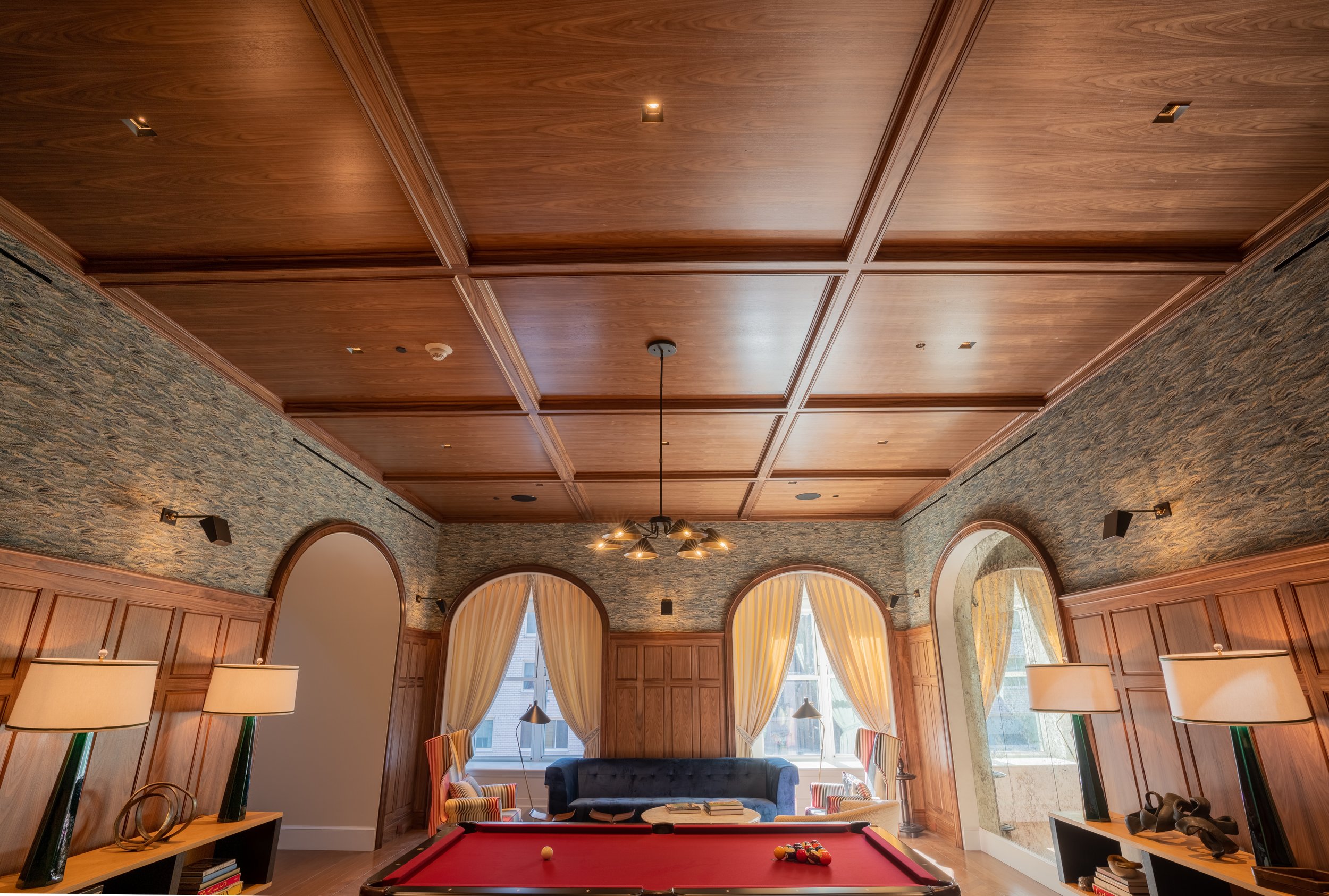 A cozy game room with a red pool table, wooden paneling on the walls, a stone-textured ceiling, large arched windows with cream curtains, a central black sofa, armchairs, and various lamps and decorative items.