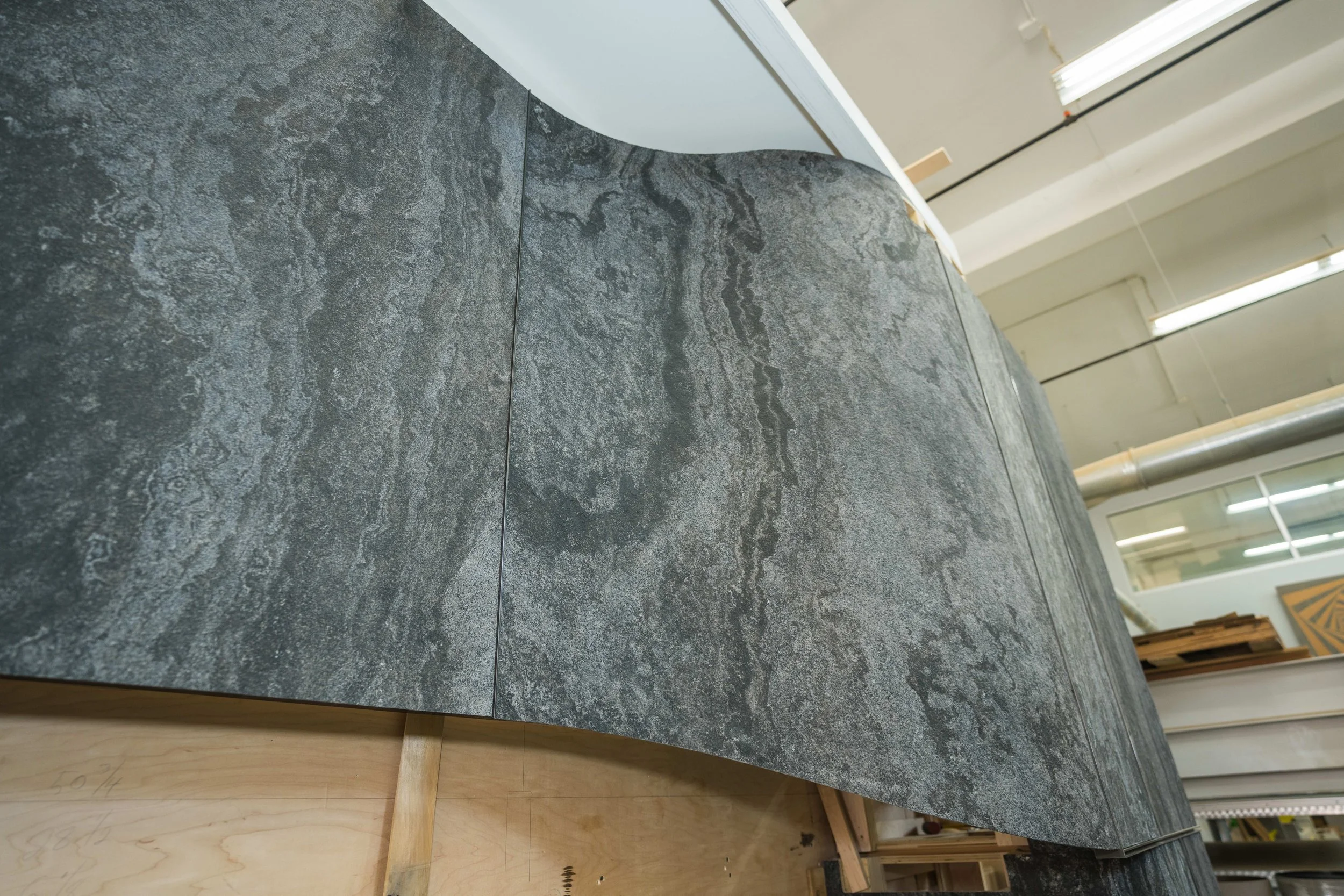 Close-up of a dark gray, textured stone counter or table with a curved edge, in a workshop or warehouse setting with fluorescent lights and various materials in the background.