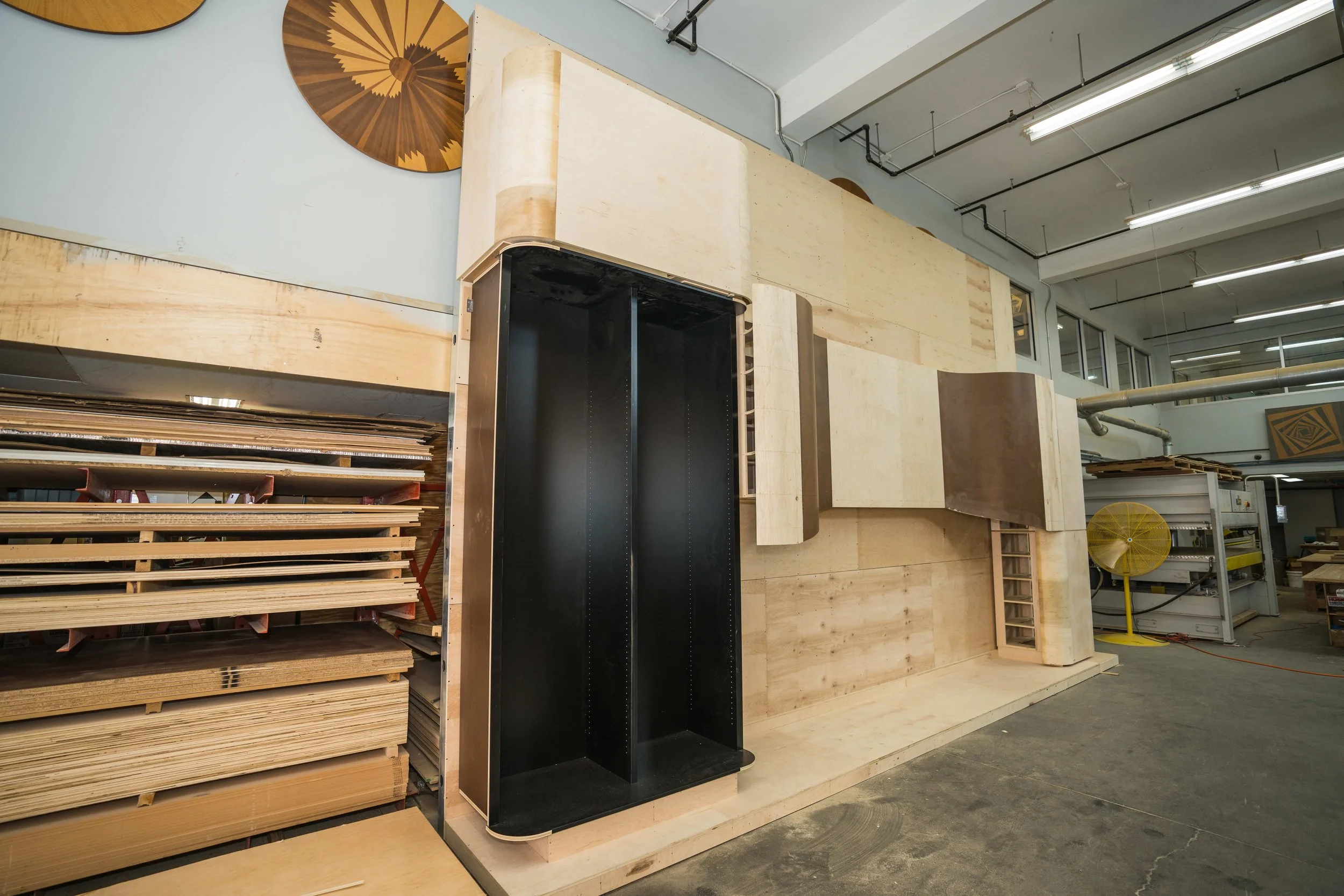 Wooden shelves and panels in a woodworking workshop, with a fan and fluorescent lights overhead.