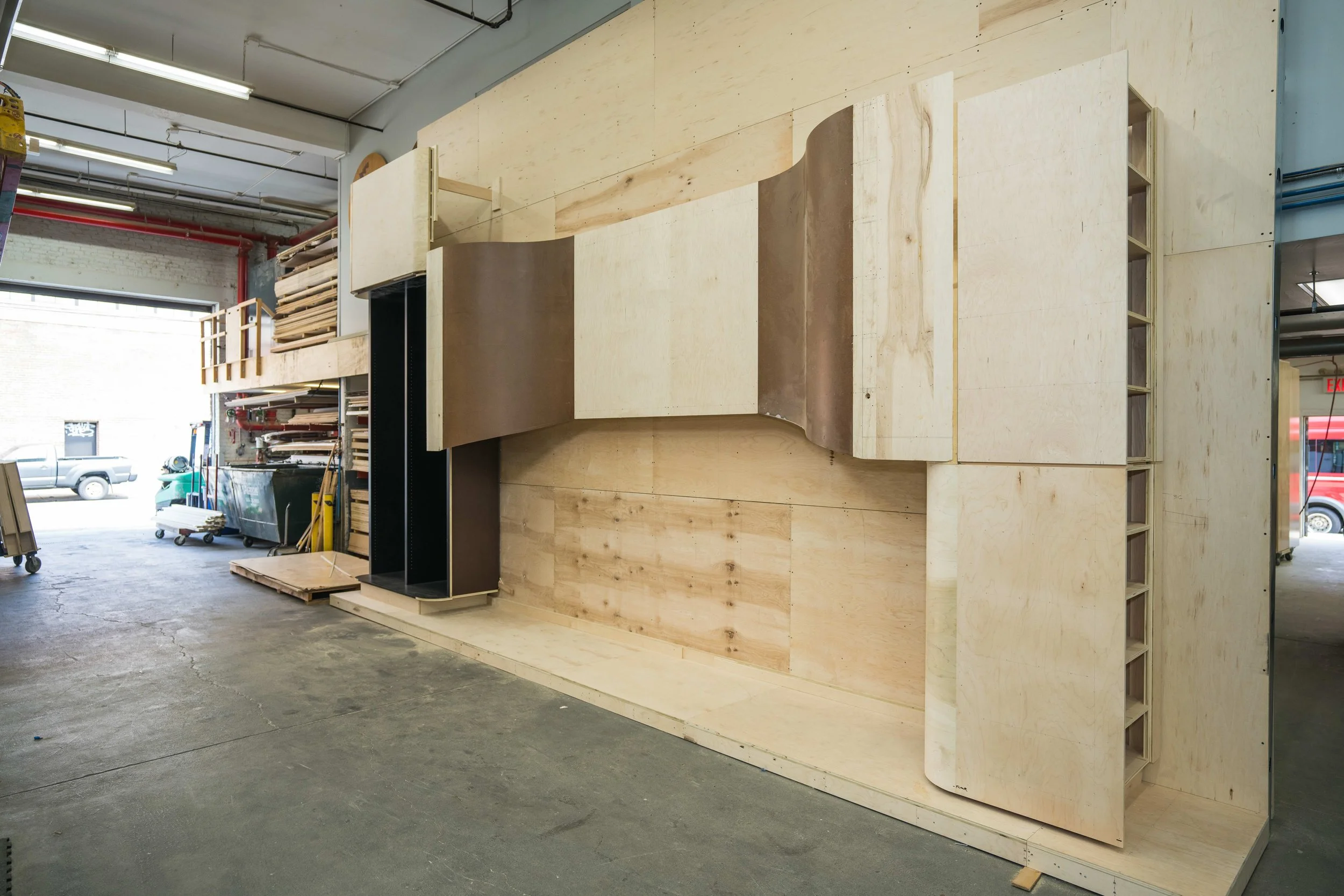 Large wooden structure or furniture piece under construction inside a workshop or warehouse, with shelves and tools around.
