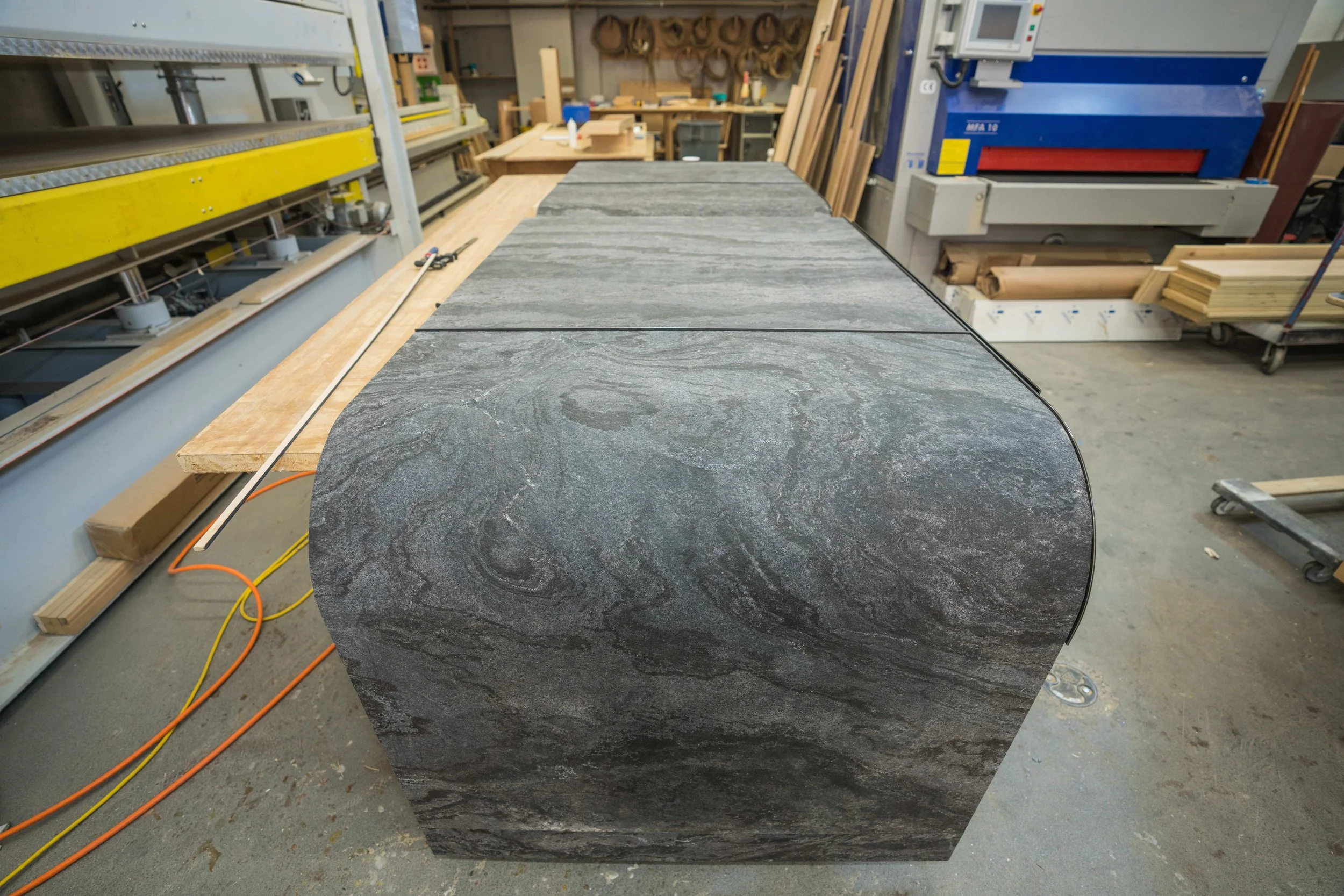A large, curved slab of dark gray stone with swirling patterns, placed on a worktable in a woodworking shop.
