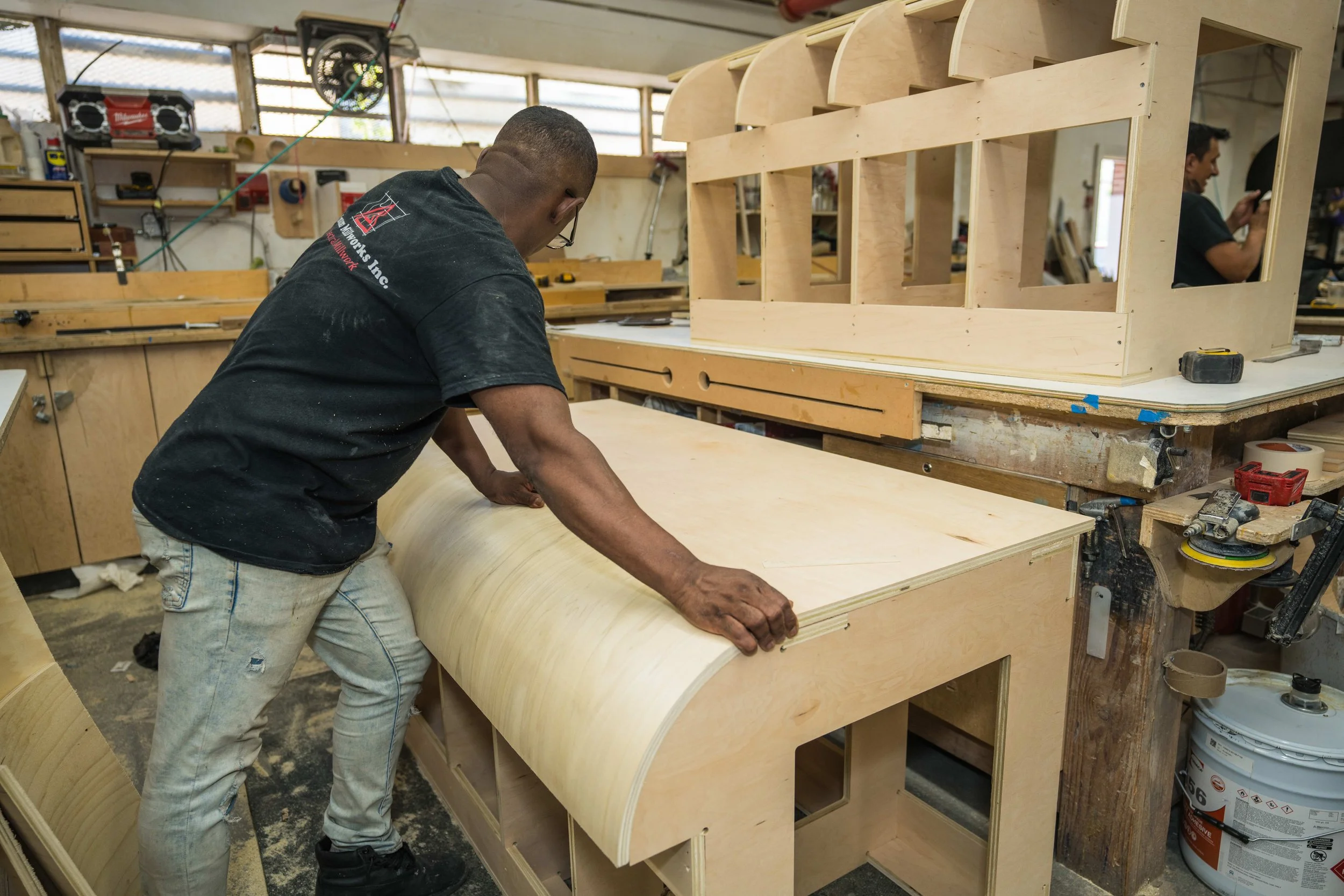 A man working on a woodworking project in a workshop, shaping and sanding plywood in front of a partially built wooden structure with multiple compartments.
