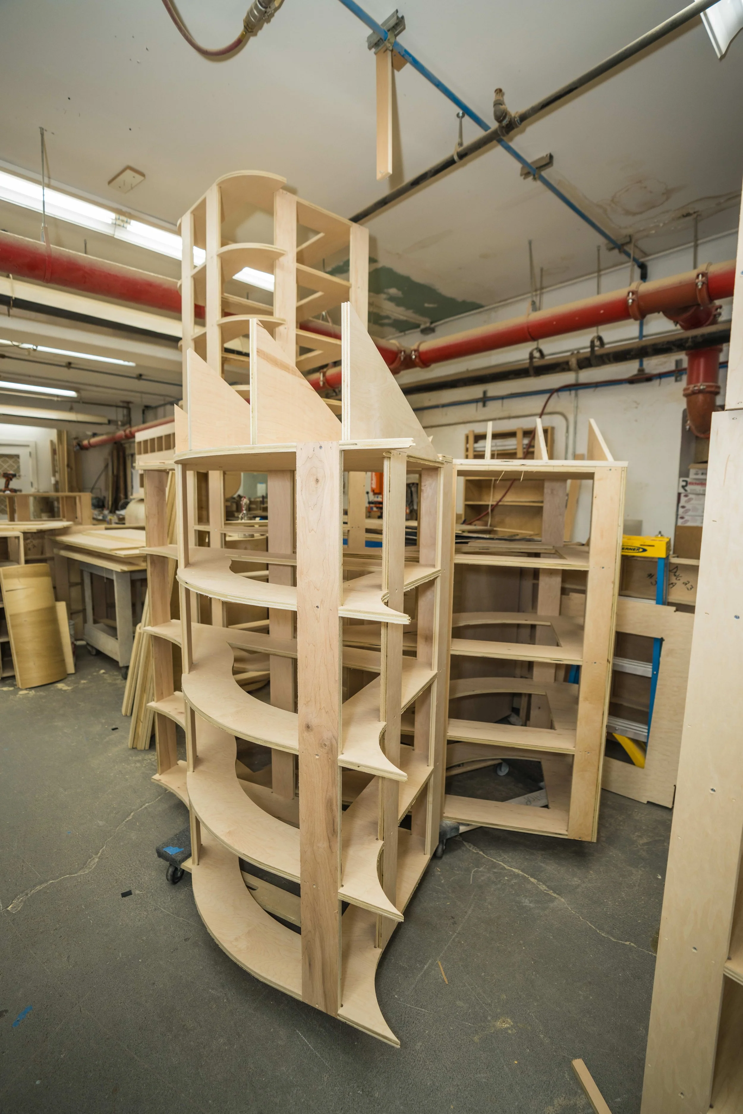 Wooden frame structure under construction in a workshop, with multiple curved shelves and vertical supports, surrounded by tools and other woodworking materials.
