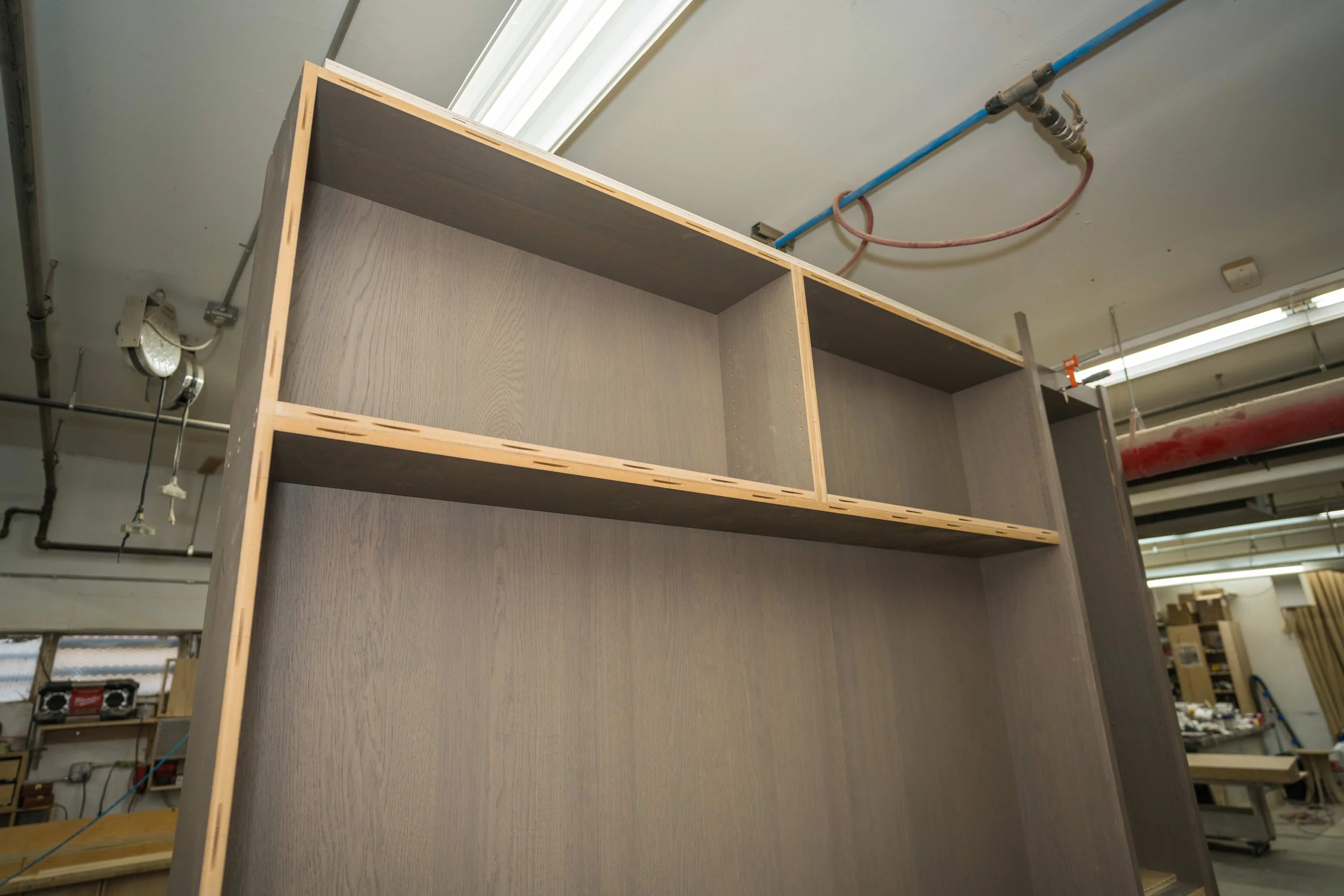 Wooden shelving unit under construction in a workshop, with visible pipes and electrical wiring on the ceiling.