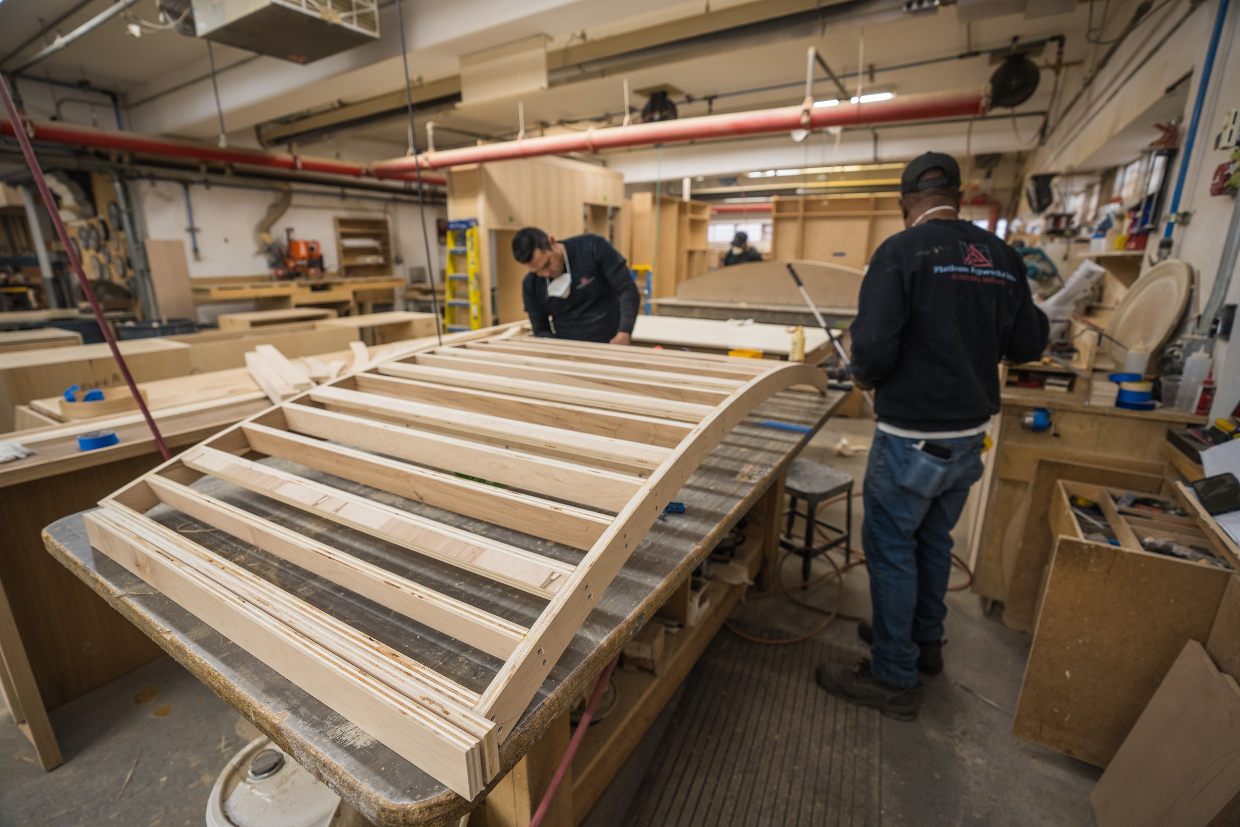Two workers in a woodworking shop constructing a curved wooden structure, with various tools and wooden parts on the workbench.