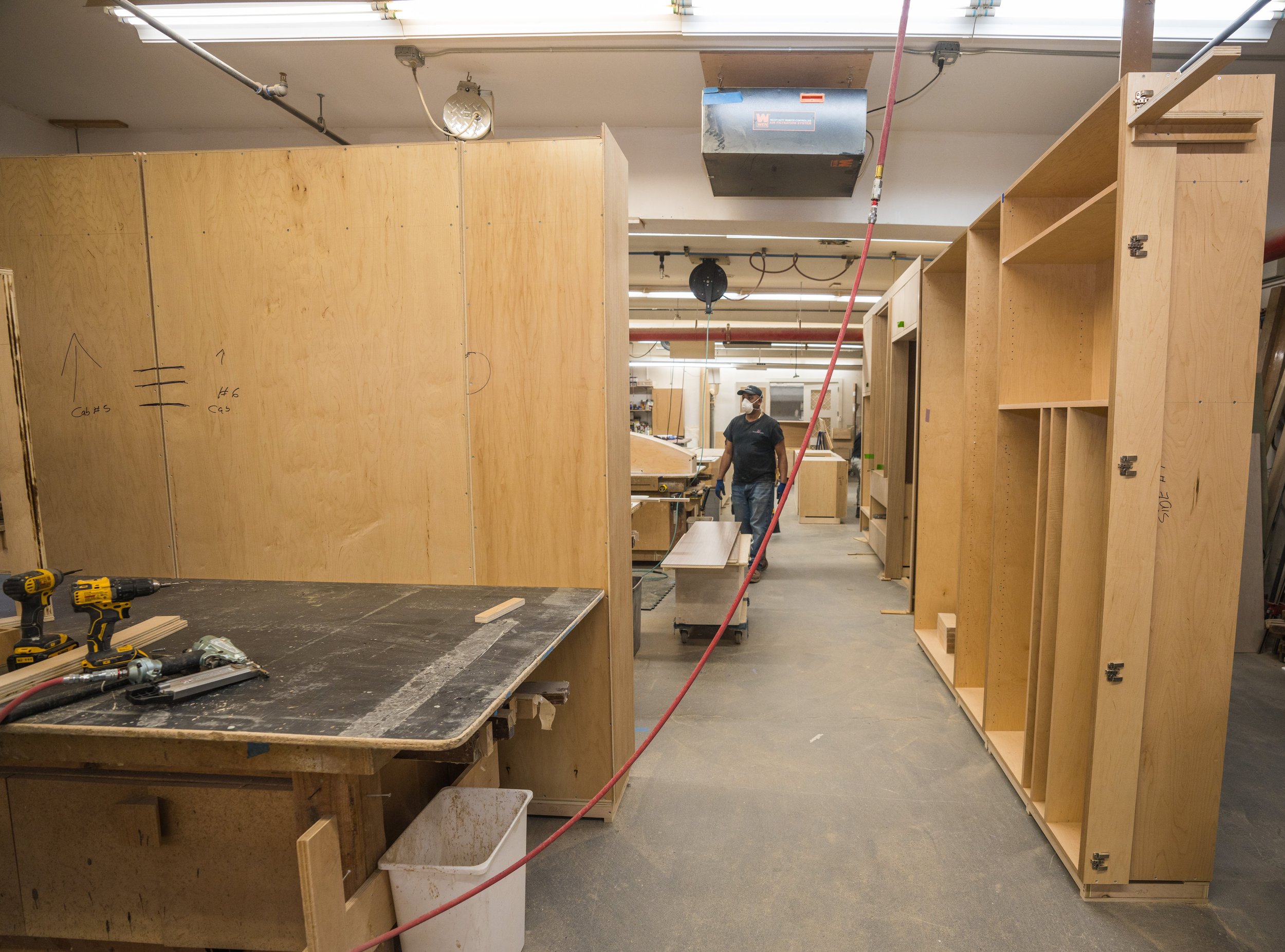 A woodworking workshop with unfinished wooden cabinetry and shelves, tools on a workbench, and a worker in a mask and gloves walking through the space.