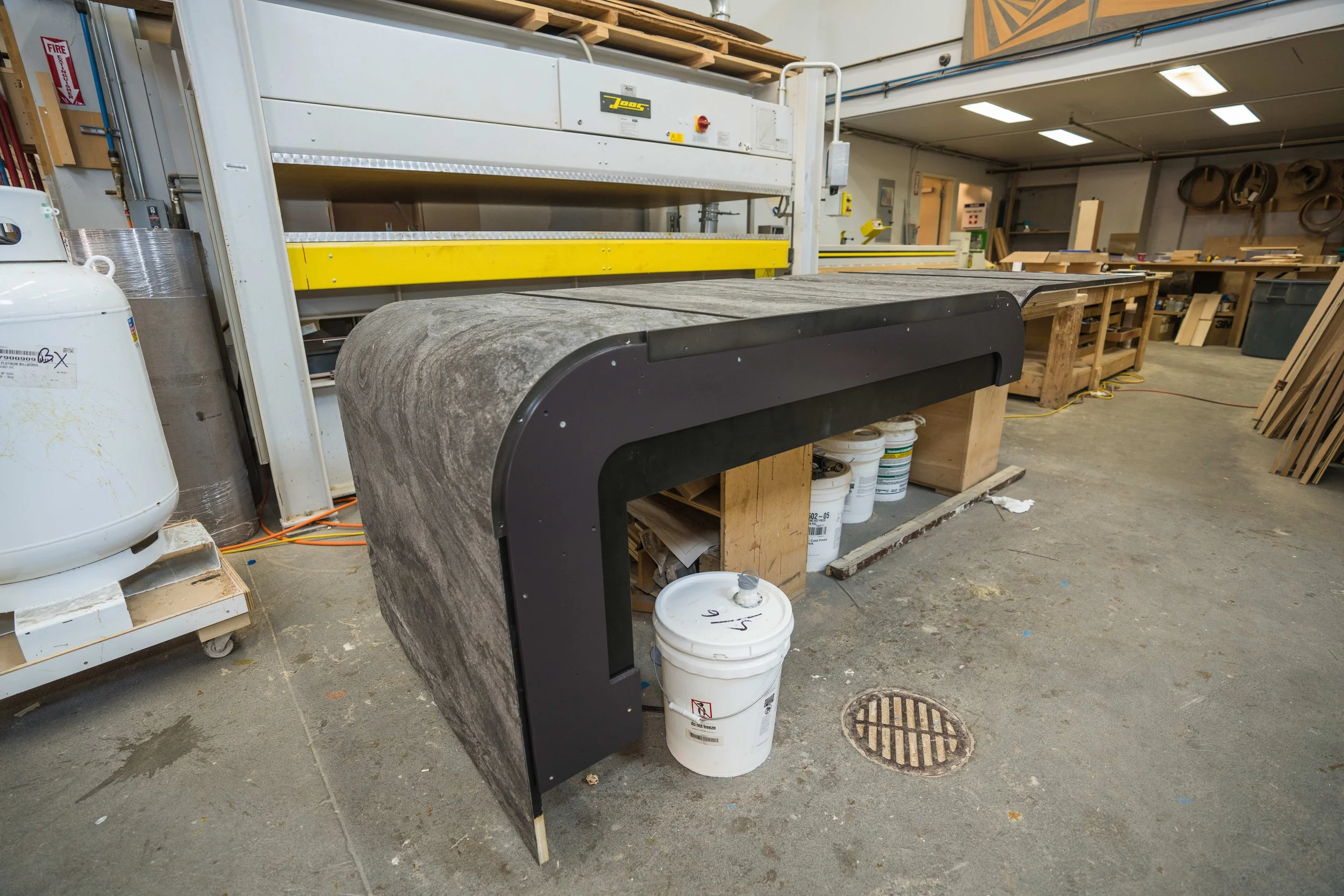 Industrial workshop with woodworking equipment, workbenches, and storage shelves, including a large granite or marble slab on a saw machine, buckets, tools, and supplies.