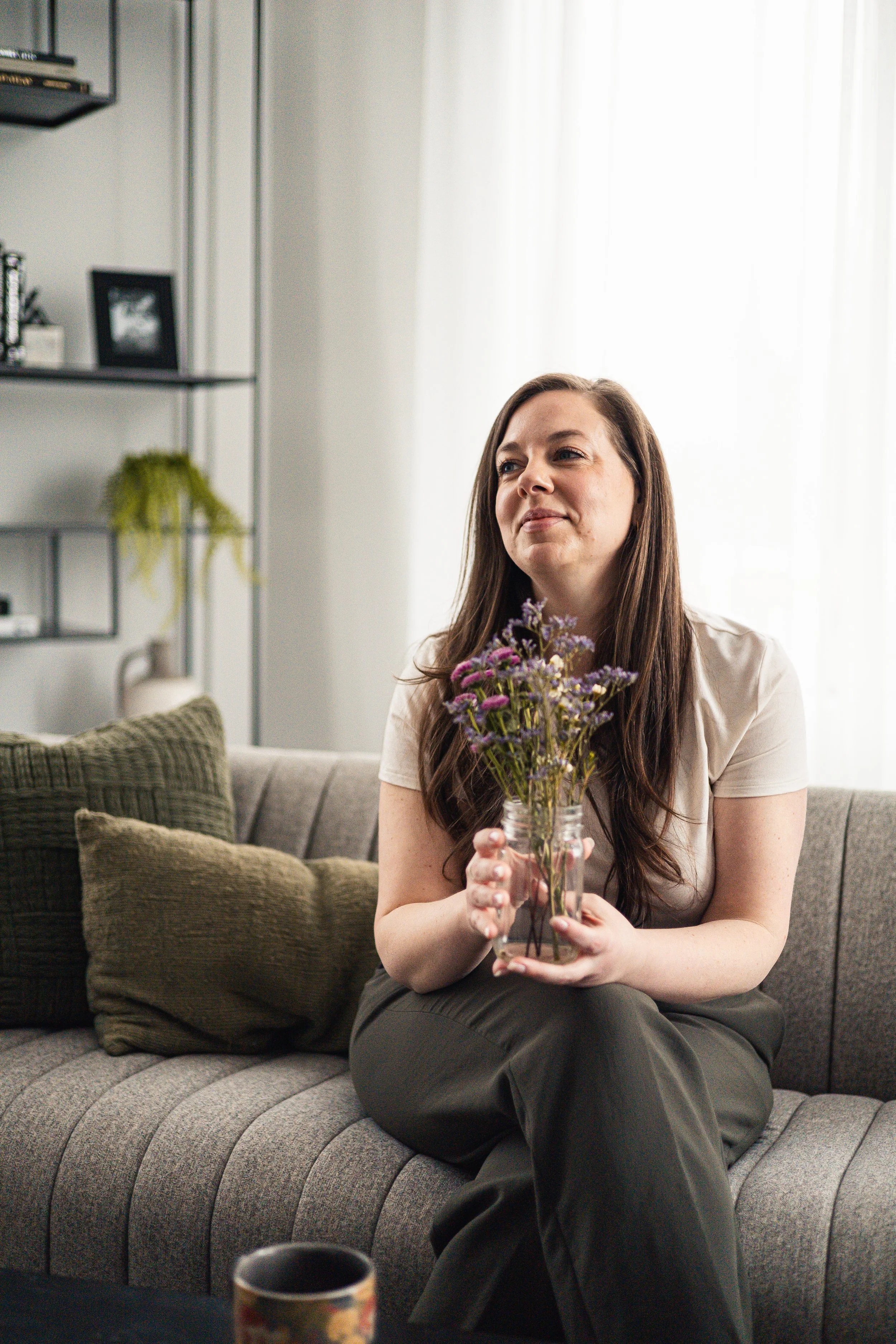 A woman with long brown hair sits on a gray couch holding a glass jar filled with purple flowers, in a well-lit living room with beige curtains and decorated shelves.