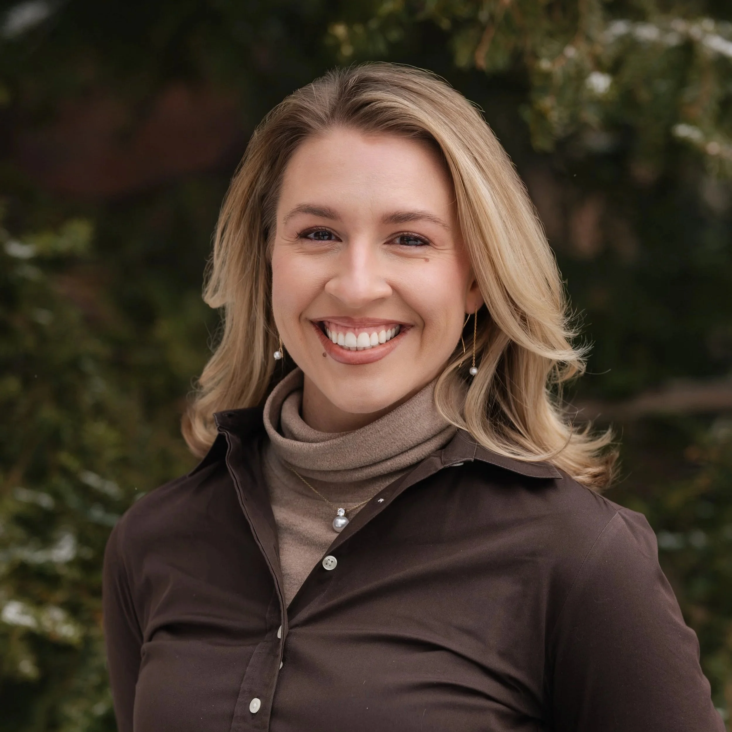 A smiling woman with blonde hair, wearing pearl earrings, a beige turtleneck, and a dark button-up shirt, outdoors with blurred trees in the background.