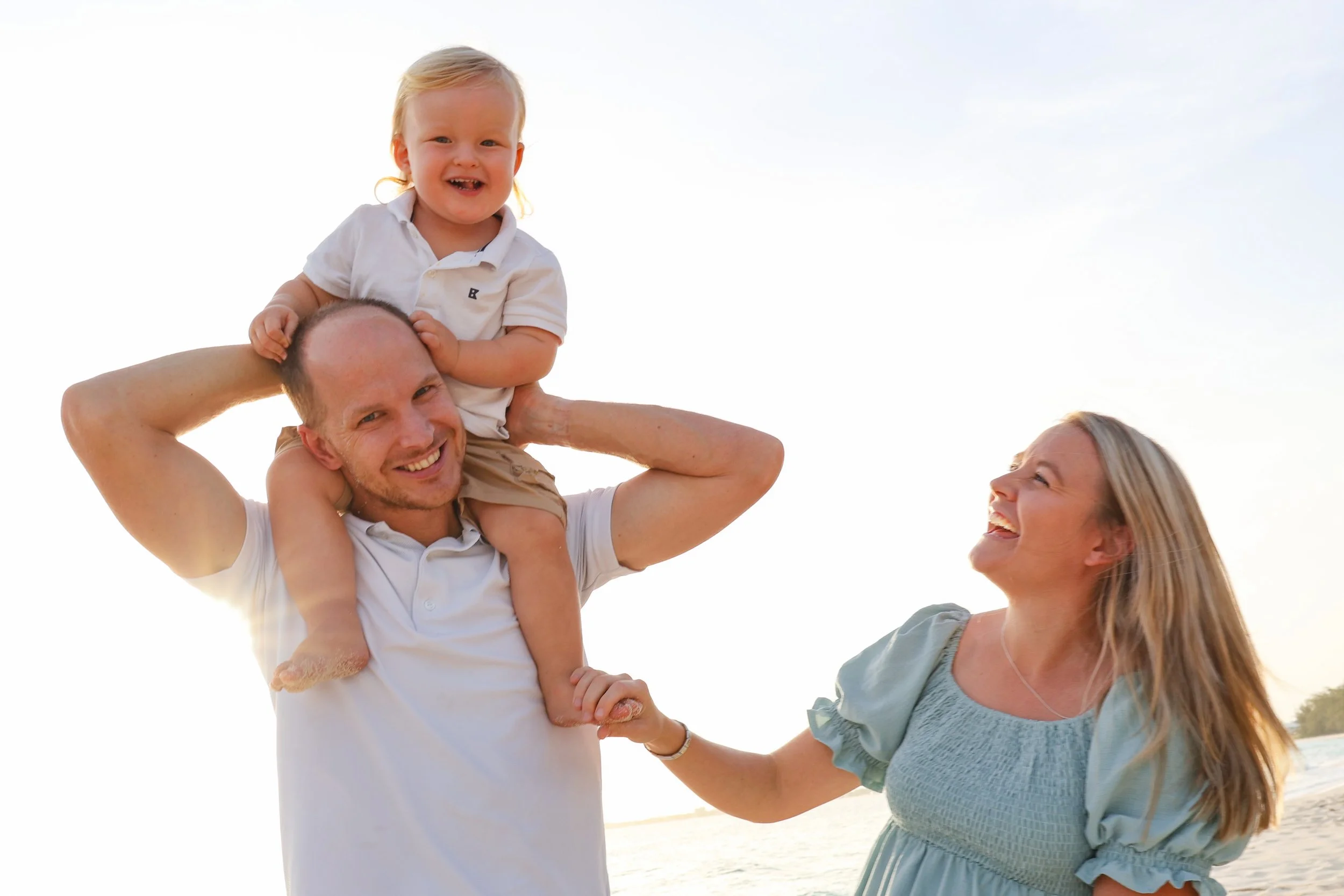 A family at the beach enjoying a sunny day. The father is holding a young child on his shoulders, and the mother is smiling and reaching out to the child. The sky is clear, and the family appears happy.