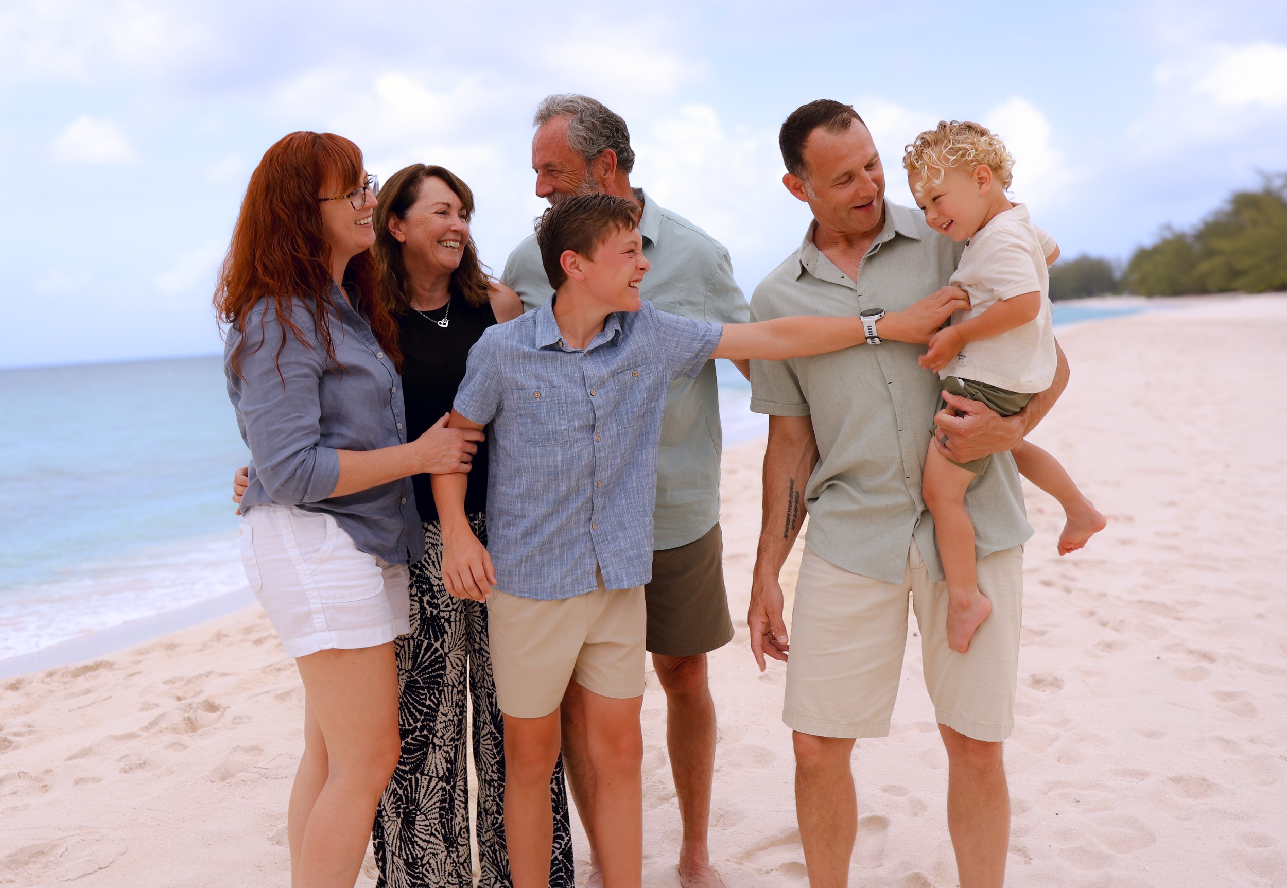 A group of seven people, including children, standing on a beach, smiling, and engaging with each other during daytime.