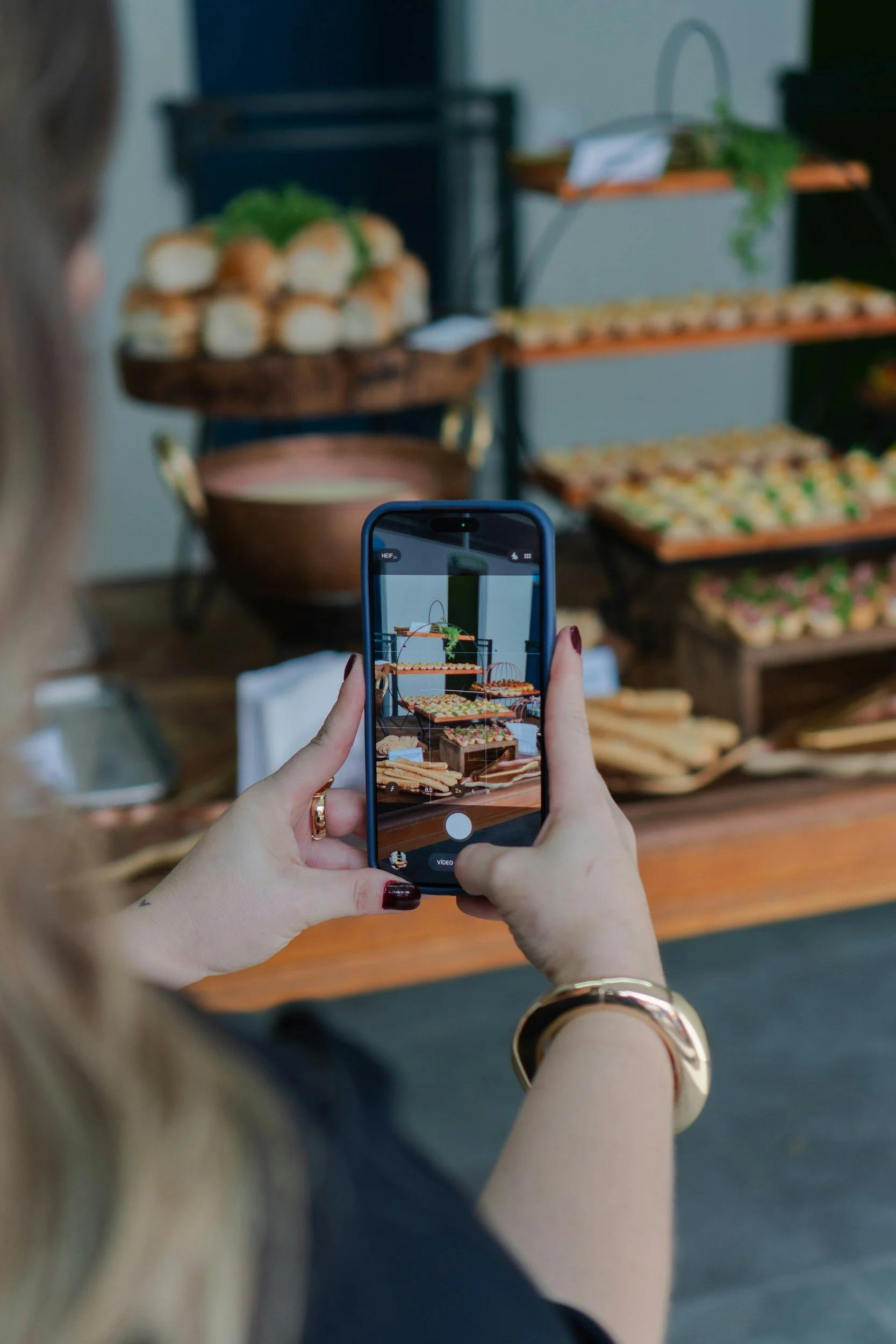 A woman taking a photo of a display of assorted finger foods and desserts at an event with her smartphone.