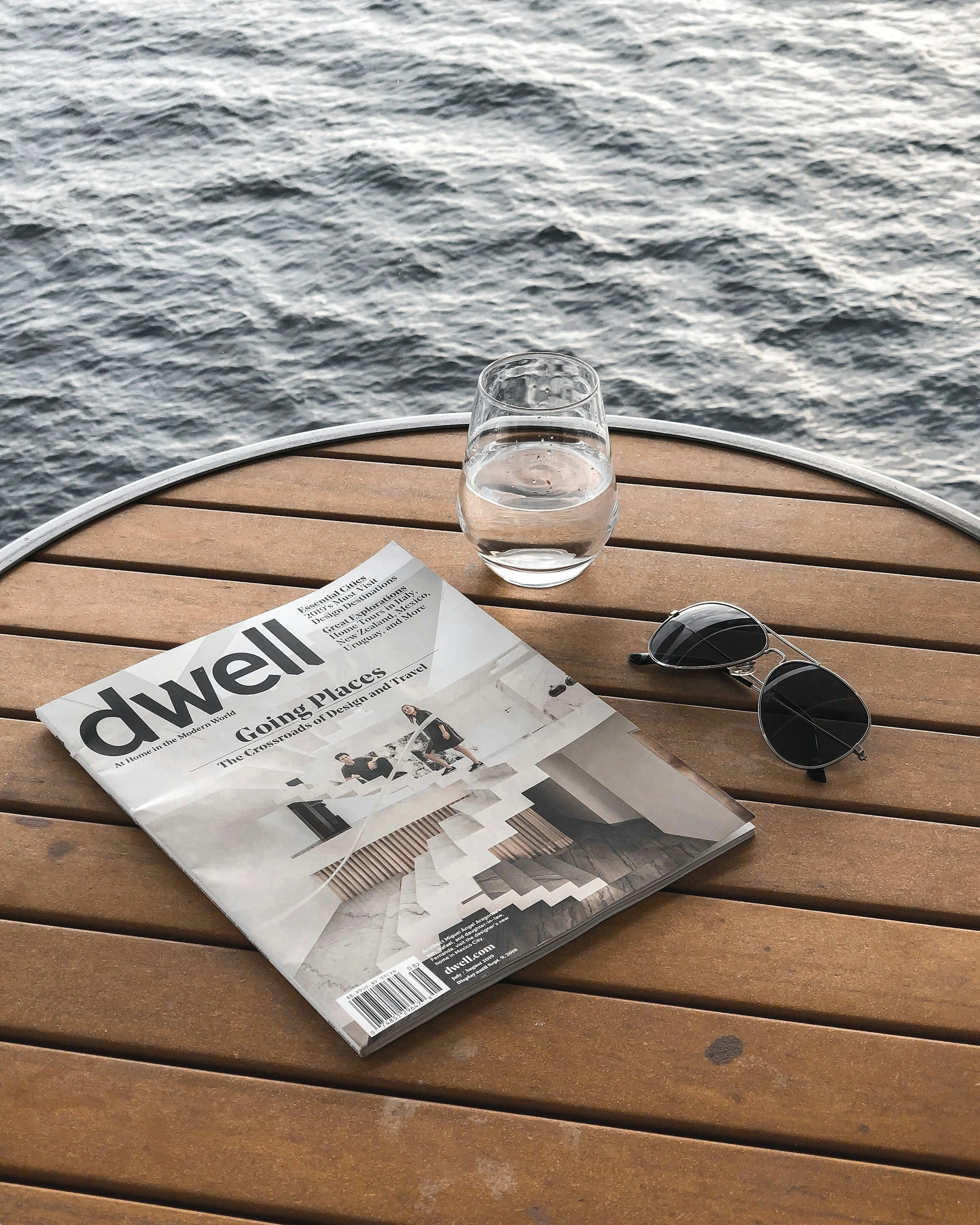 A wooden table with a glass of water, sunglasses, and a partially visible newspaper, with water in the background.