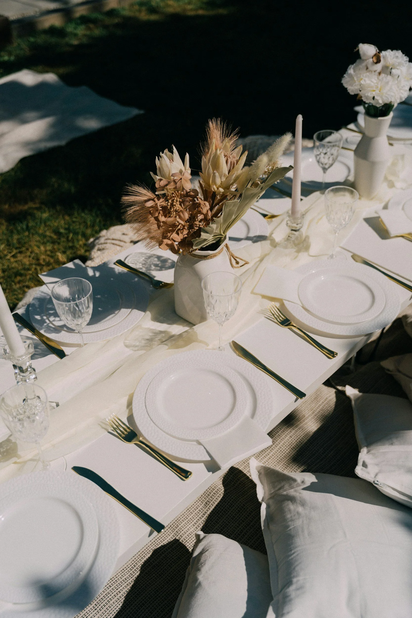 A table set for a formal outdoor event with white plates, gold flatware, and crystal glasses. There are two vases with white and pink dried flowers as centerpieces. The table is covered with a white tablecloth, and there are white napkins folded beside each plate. The setting is illuminated by sunlight, casting shadows on the table.