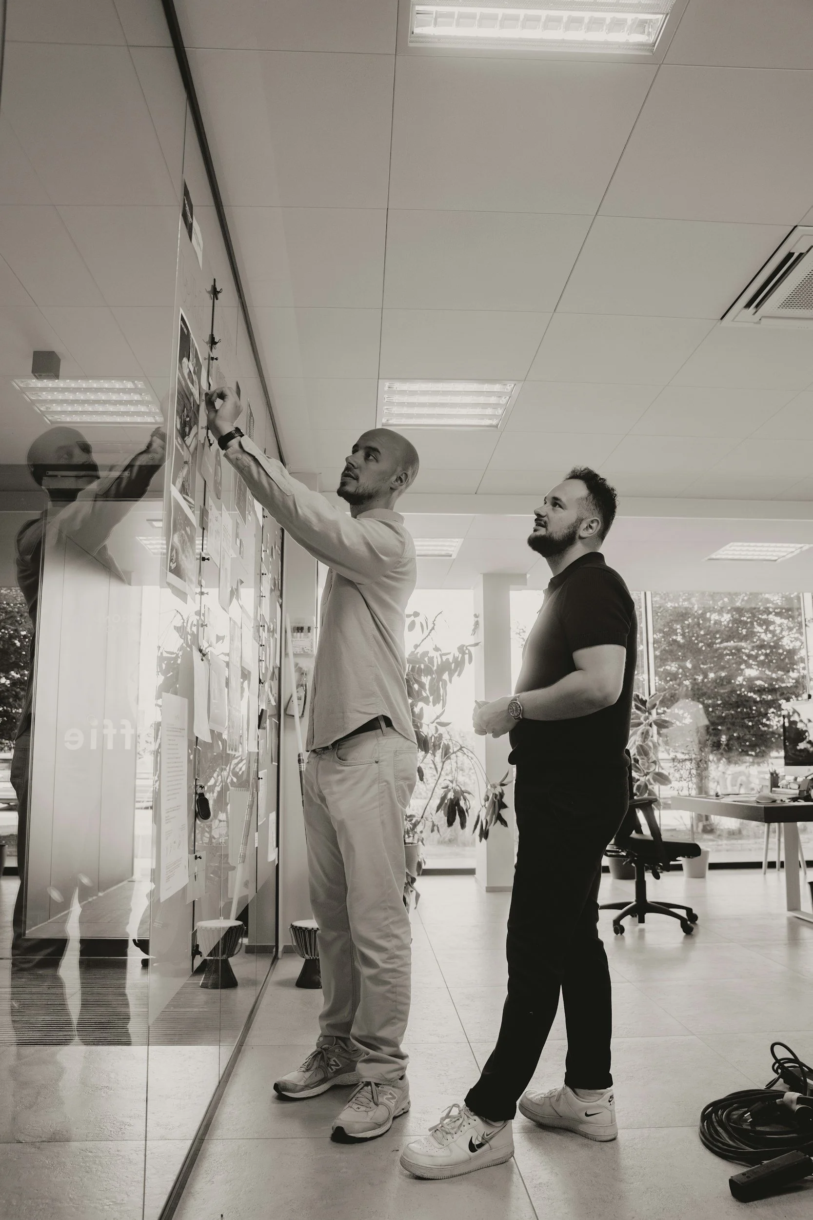 Two men standing in front of a glass wall decorated with various papers, engaging in a discussion or presentation in an office environment. One man is pointing at the wall, while the other watches attentively. The room has a ceiling with fluorescent lights and some office furniture in the background.